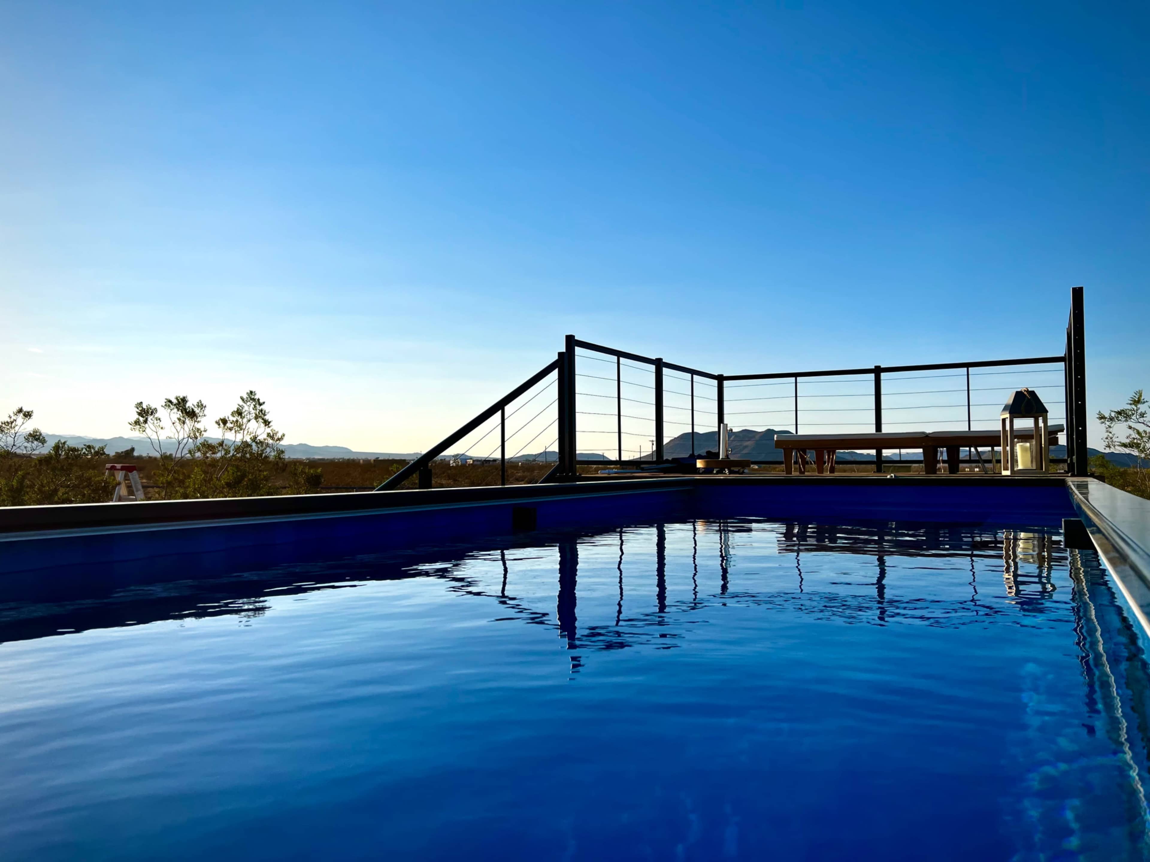 A clear, blue swimming pool reflects the sky, with a wooden deck and railing in the background overlooking a mountainous landscape.
