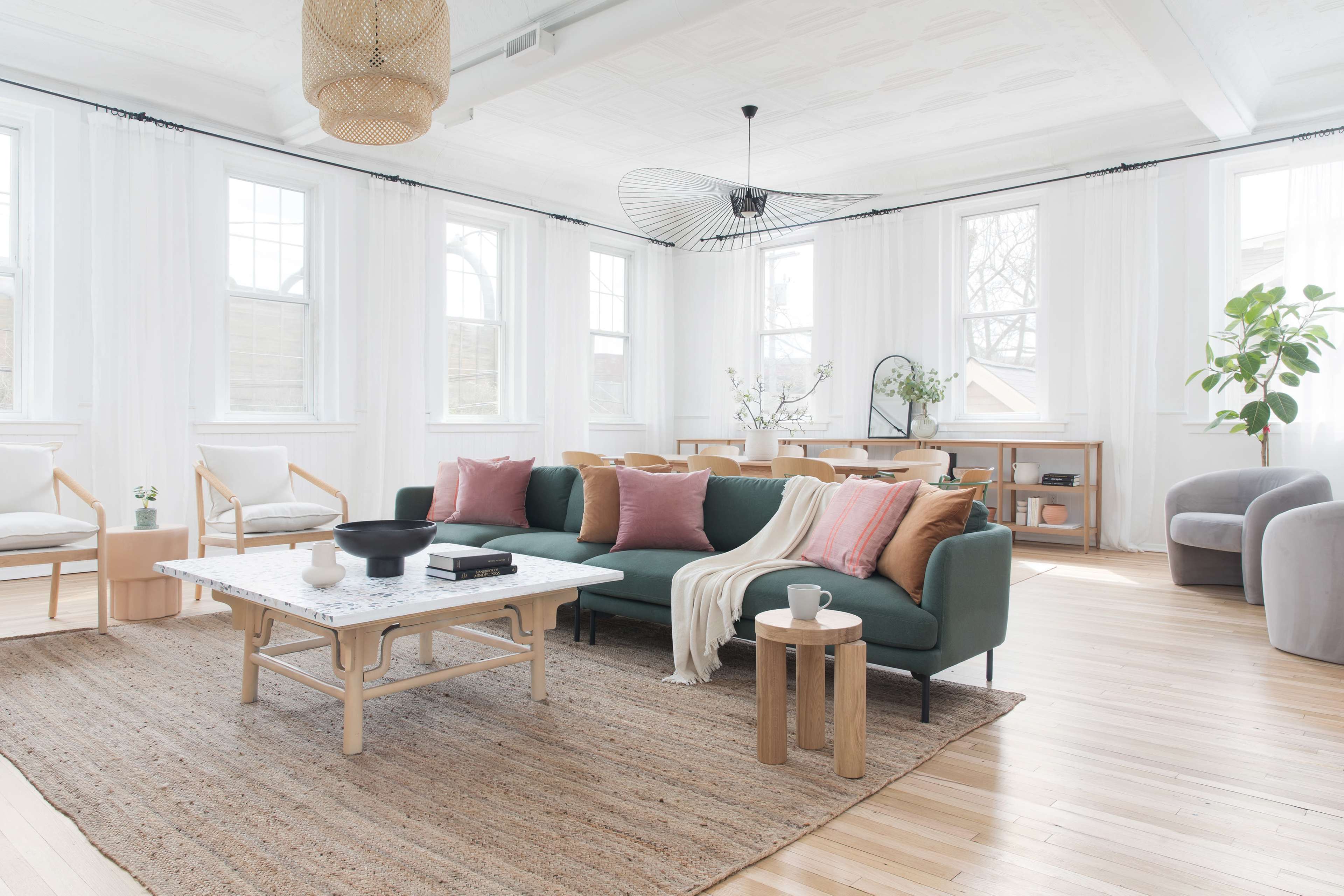 A spacious living room features a green sofa with pillows, a white coffee table, natural wood furnishings, and large windows letting in ample light.