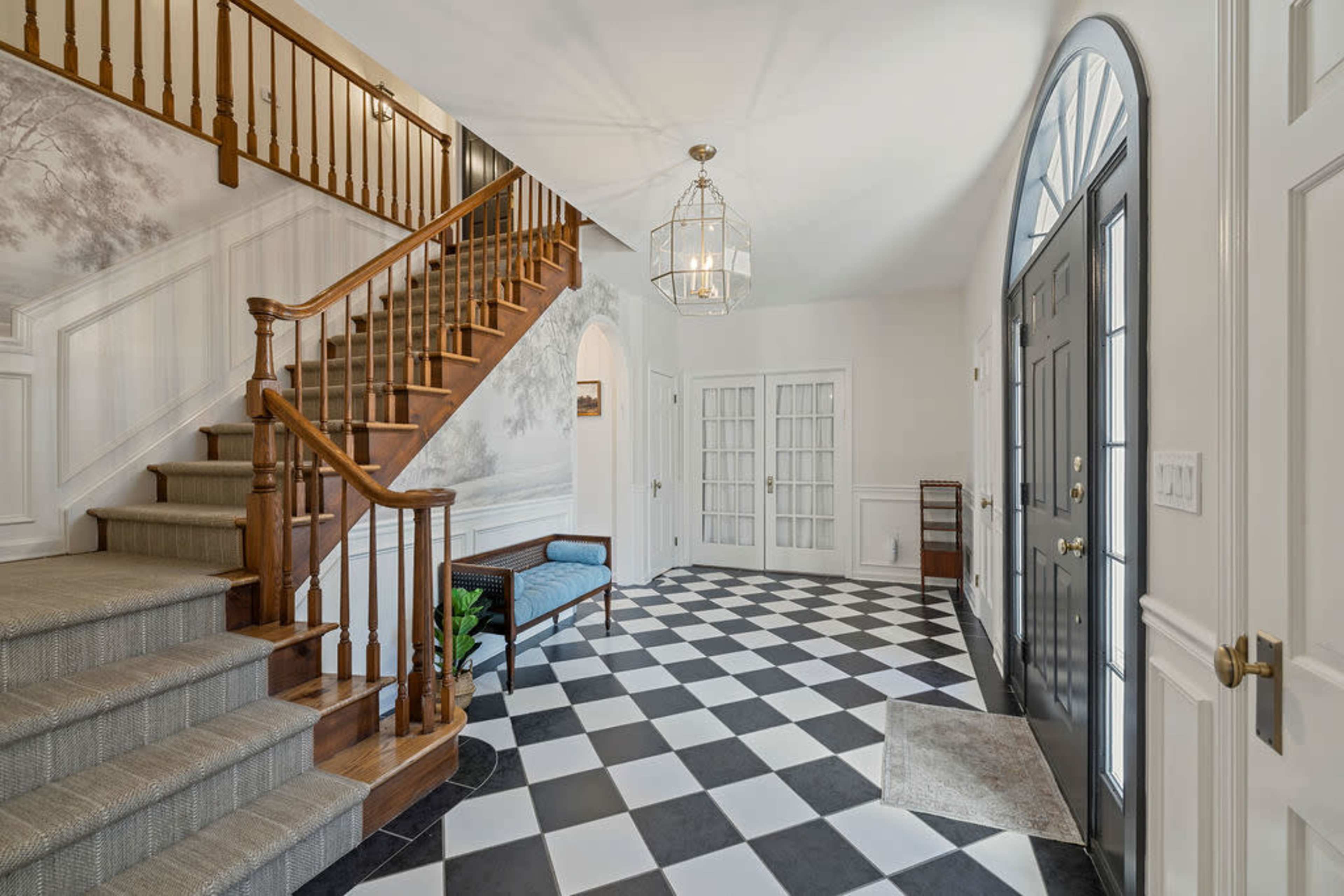 The image showcases a spacious entrance hall featuring a wooden staircase, black and white checkered floor, and a decorative light fixture.