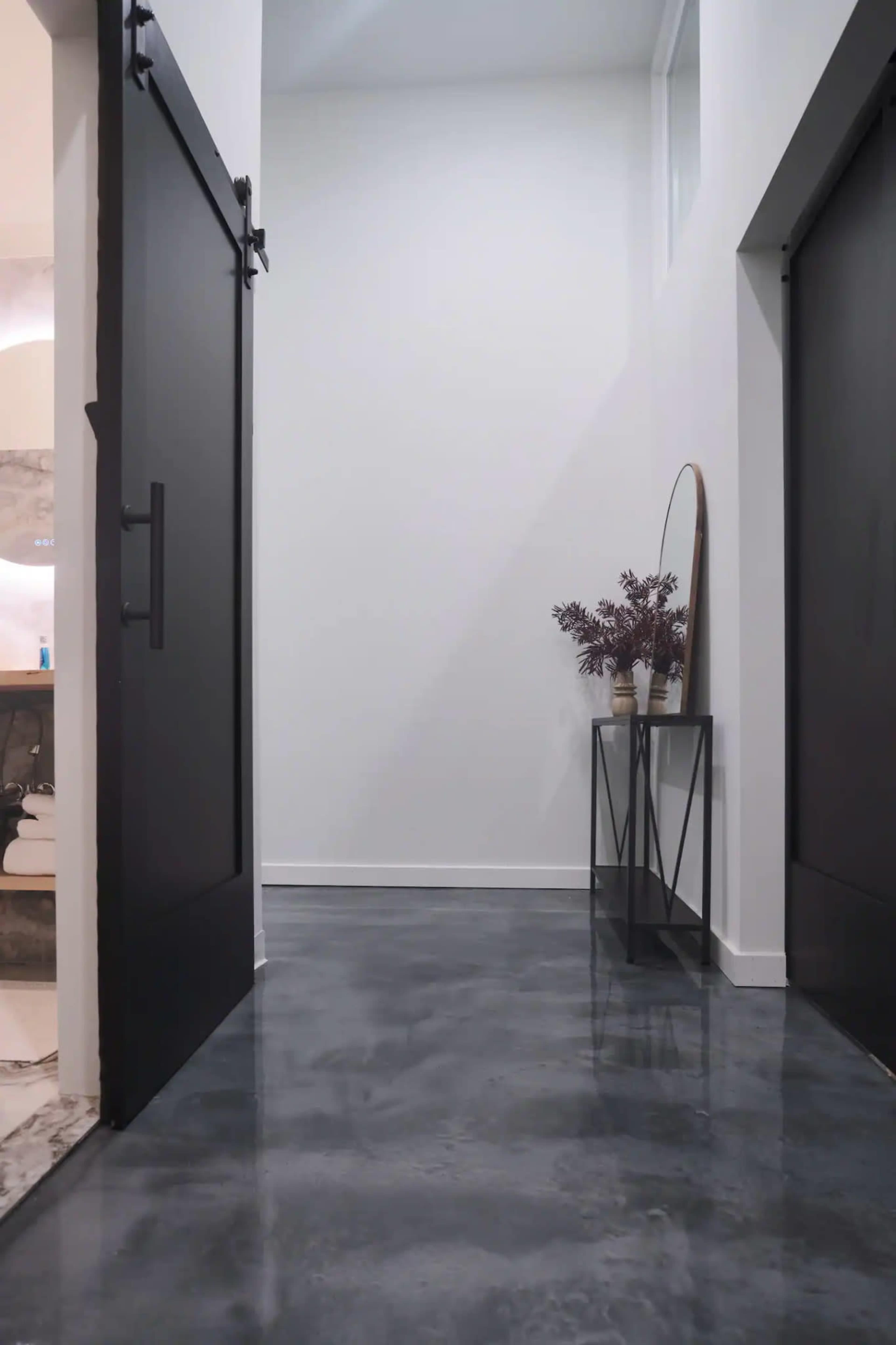 A hallway features two black sliding doors, a console table with a vase of flowers, and a polished concrete floor.