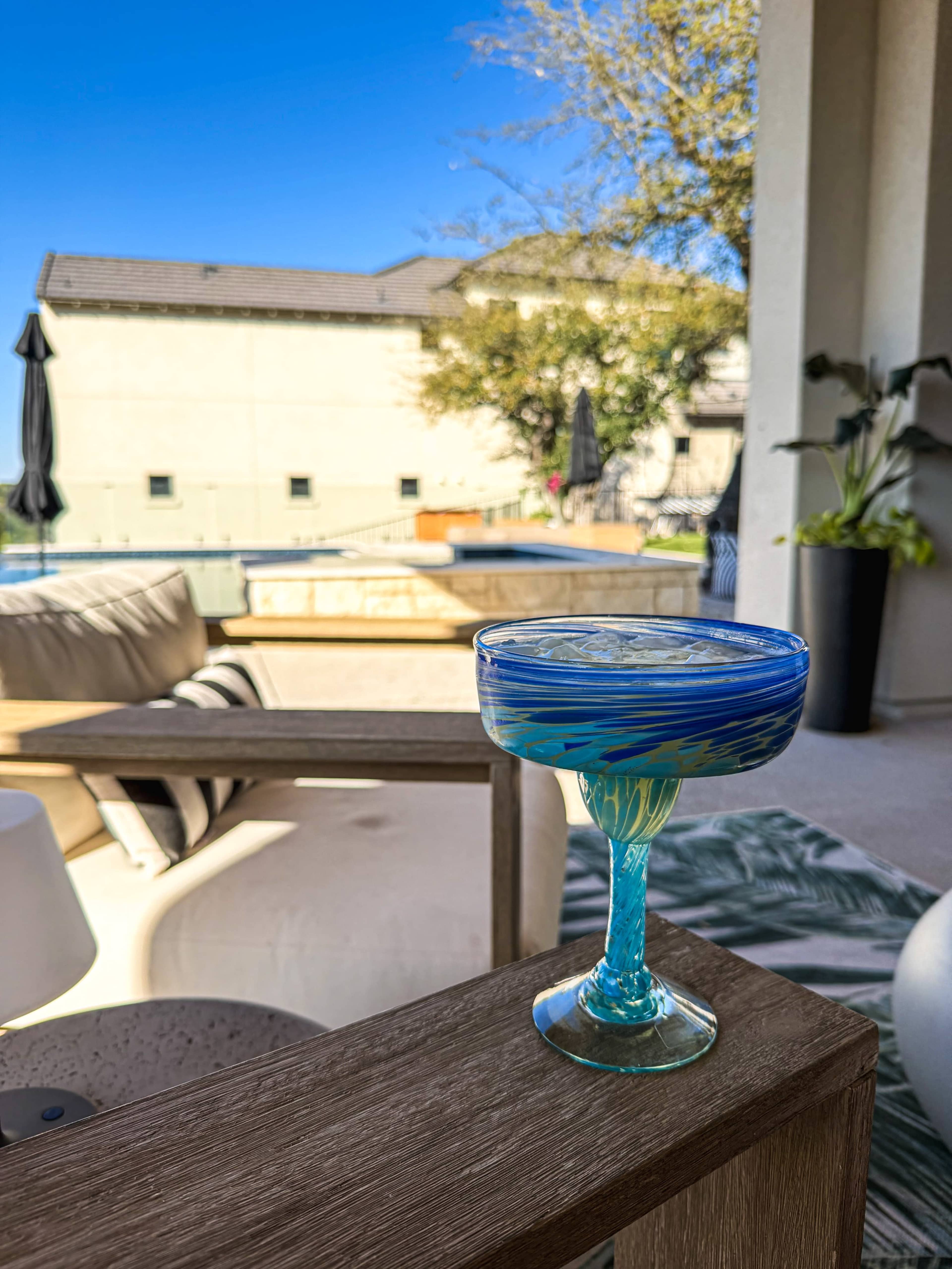 A blue cocktail sits on a wooden table in an outdoor seating area overlooking a pool and landscaped patio.