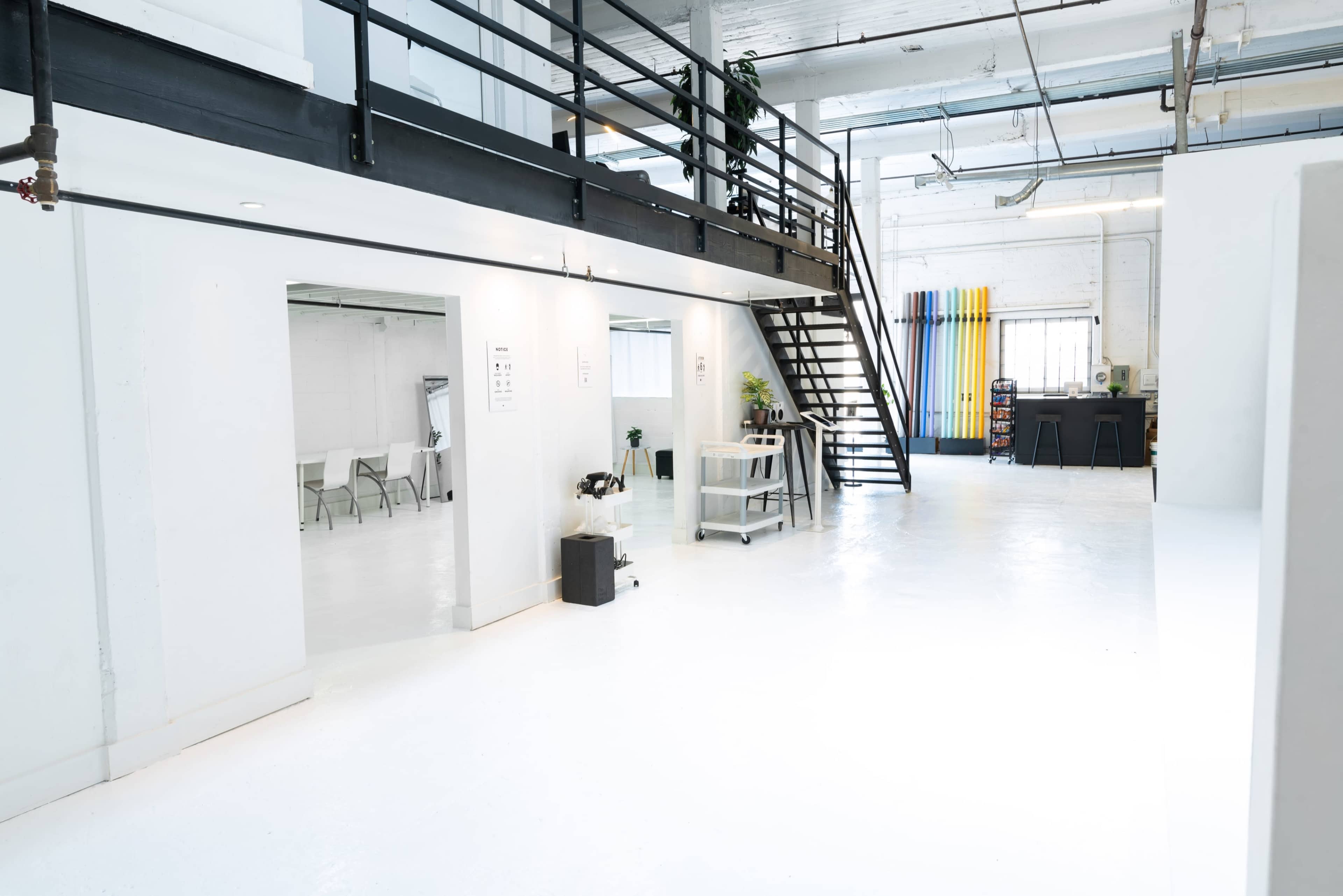A spacious, white-walled interior with a staircase on the left, leading to a mezzanine, and multiple rooms on the right, featuring simple furniture and colorful backdrop rolls in the background.
