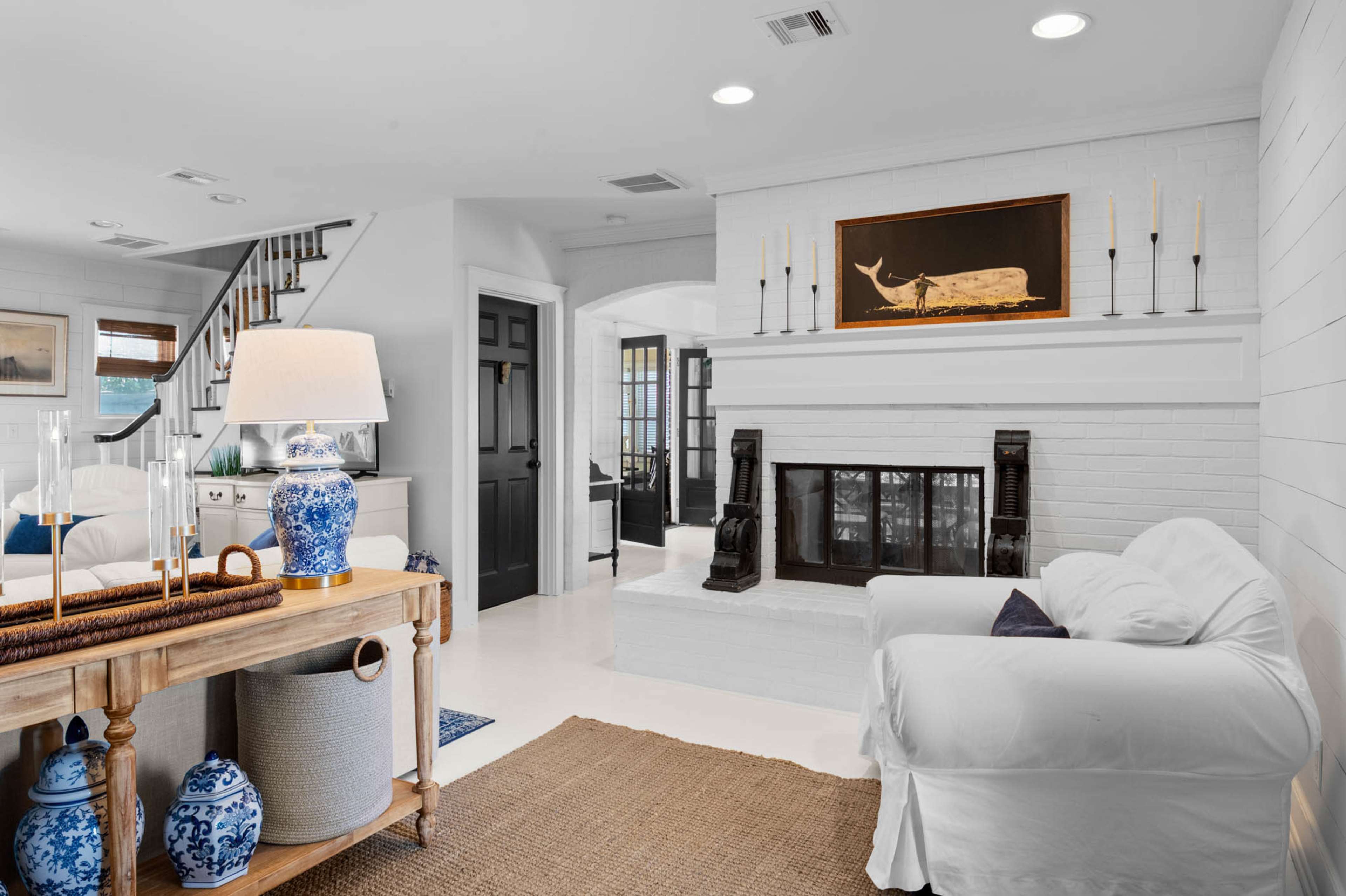 The image shows a bright living room with a white brick fireplace, a light-colored sofa, and decorative blue and white ceramic pieces on a wooden console table.