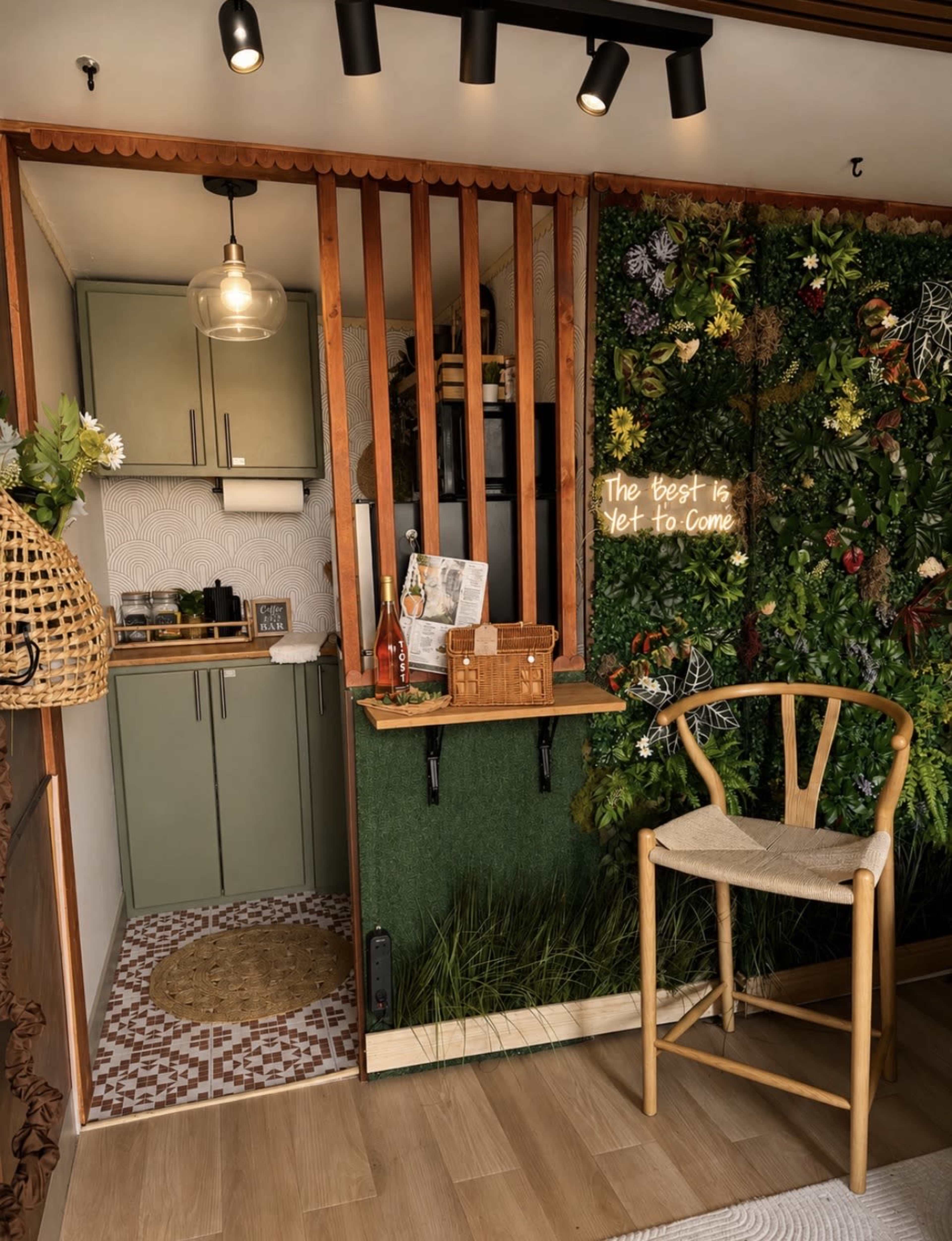 The image shows a cozy interior space featuring a kitchenette with green cabinets, a decorative wall covered in a variety of plants, and a wooden chair beside a small table.