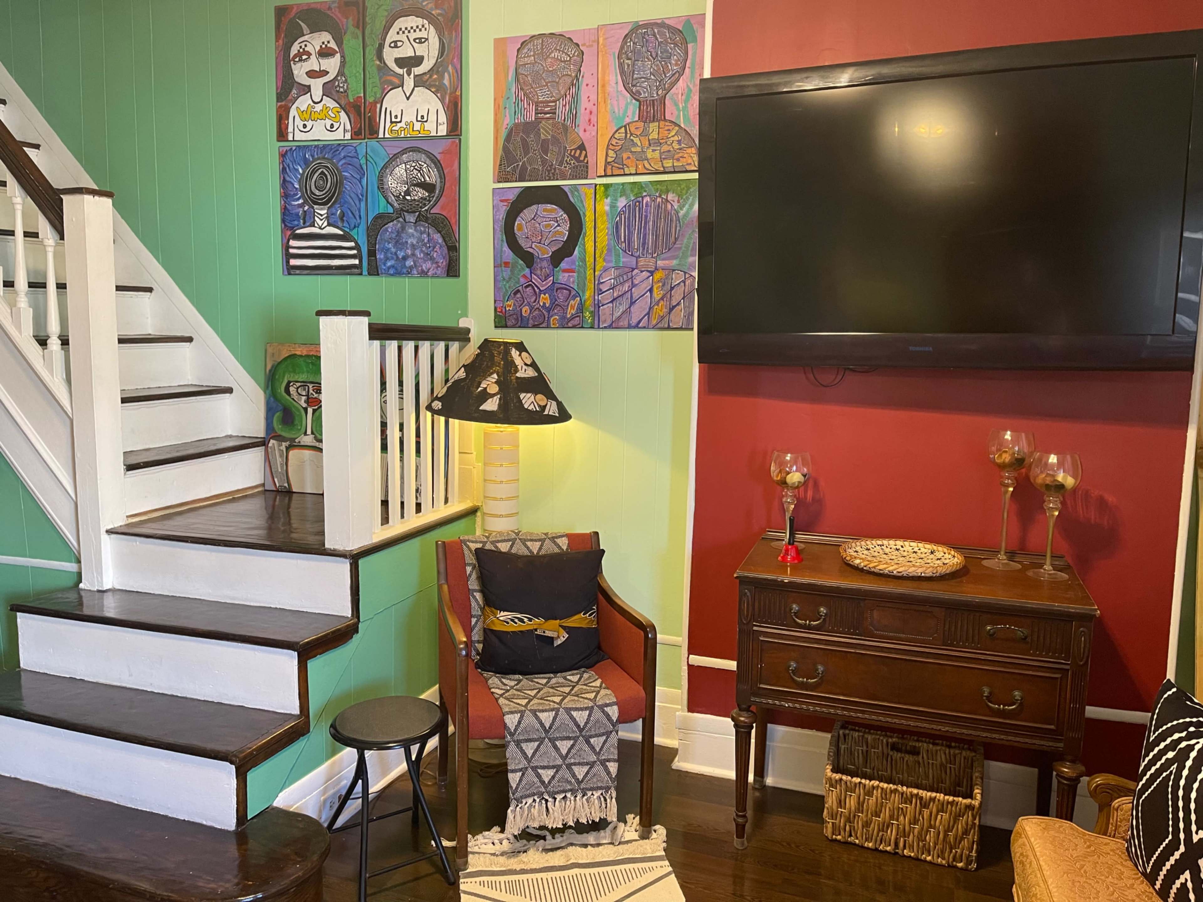 The scene shows a living space with a staircase, a wall decorated with colorful artwork, a television mounted on a red wall, and various pieces of furniture including a chair, lamp, and a wooden dresser.