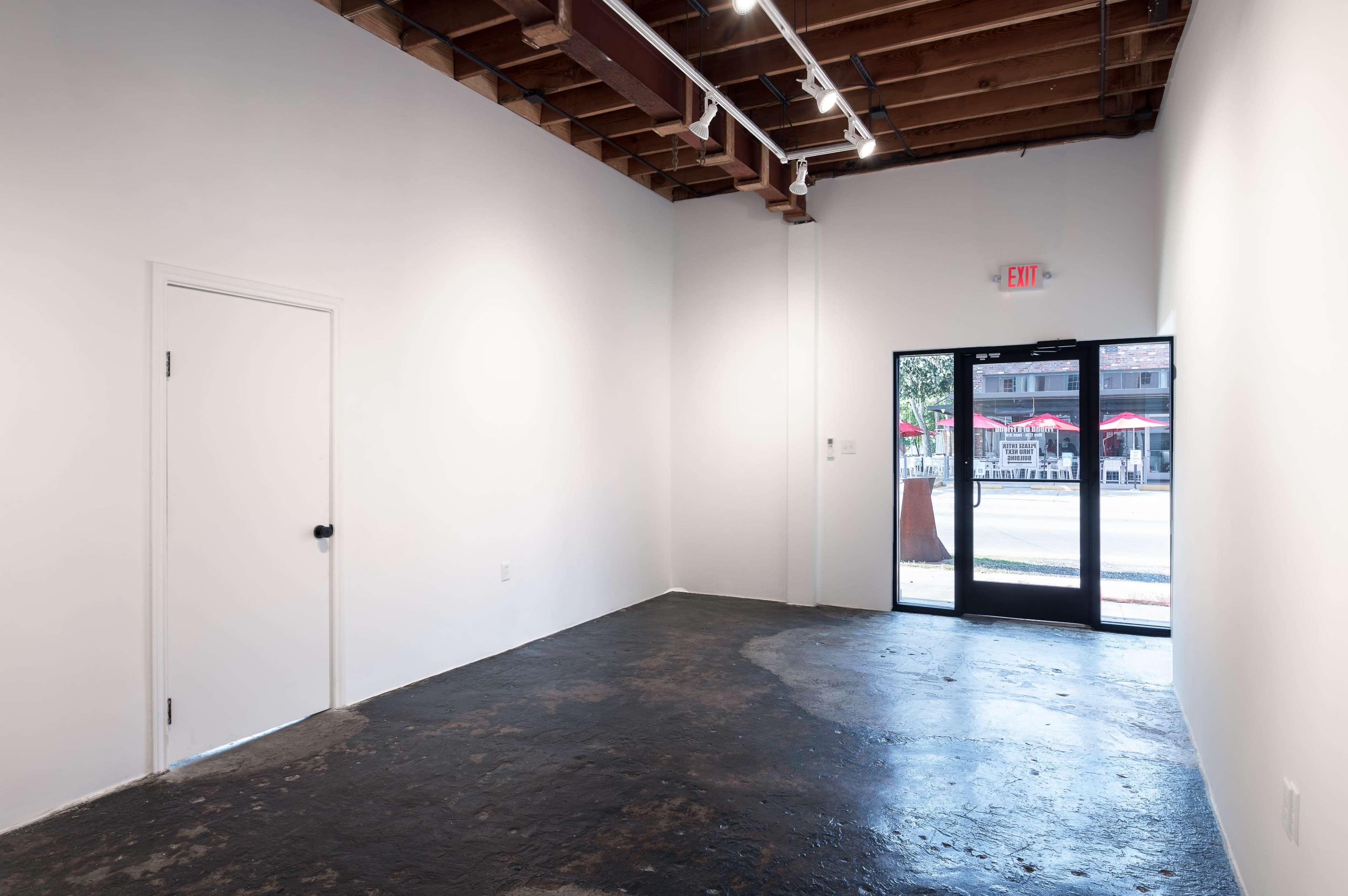 A minimalist art gallery interior with bare white walls, a dark floor, and an entrance door leading to an outdoor area with visible seating.