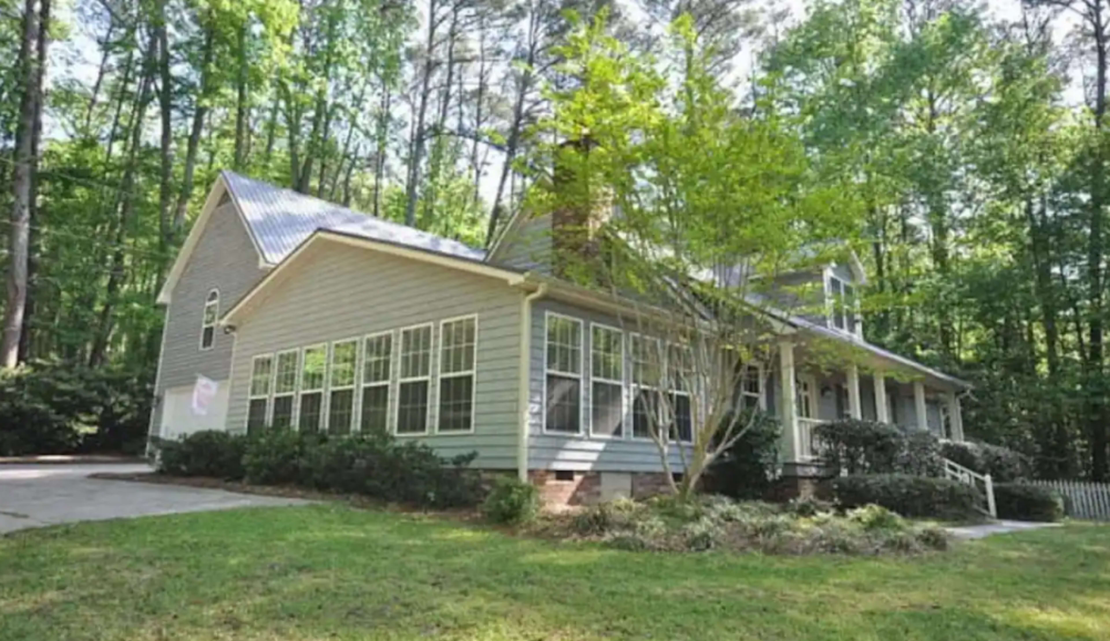 A single-story gray house with a front porch and numerous windows is surrounded by trees in a wooded area.