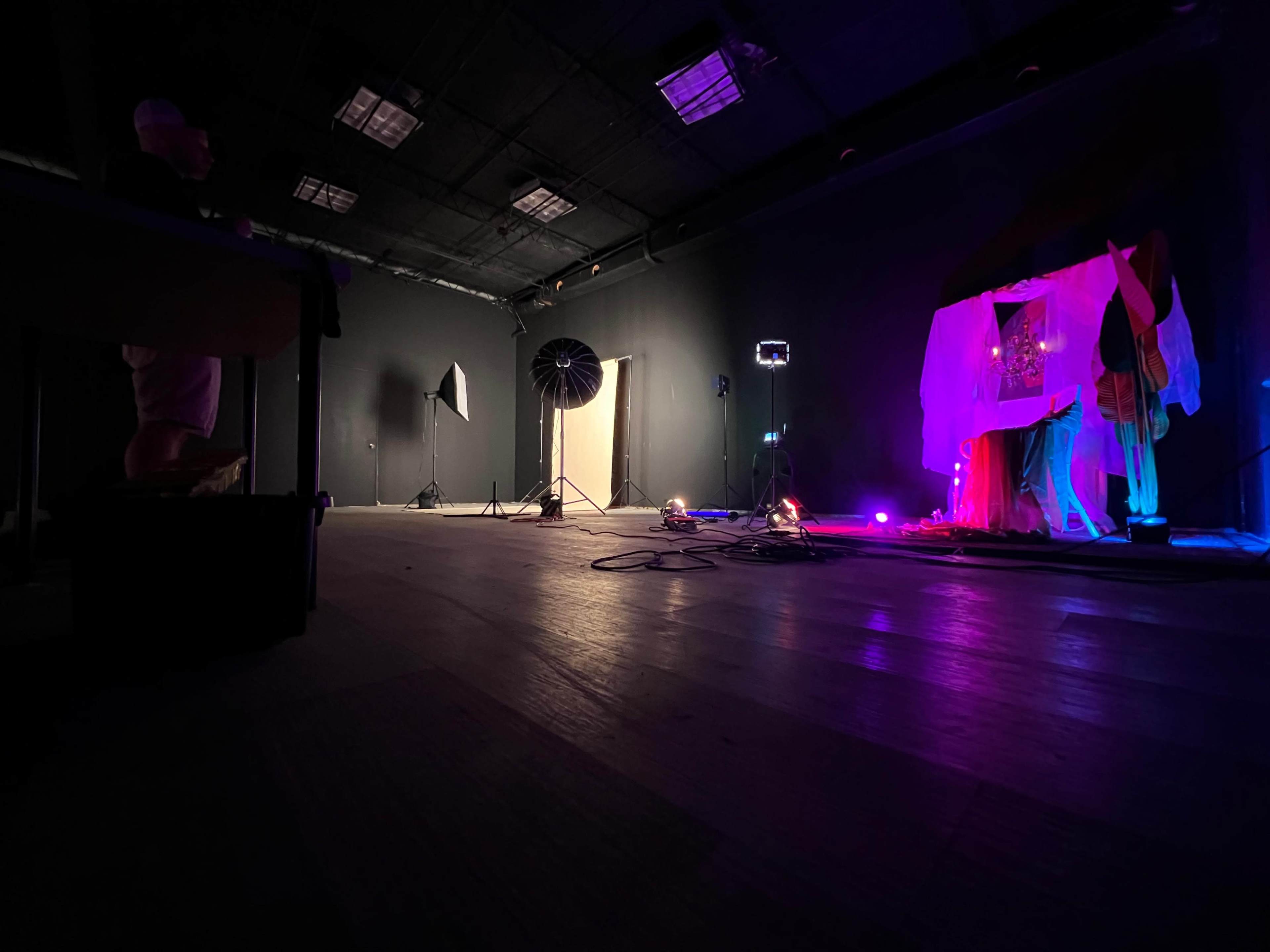 The image shows a dimly lit studio space with various lighting equipment, including a softbox and colored lights, set up for a photography session.