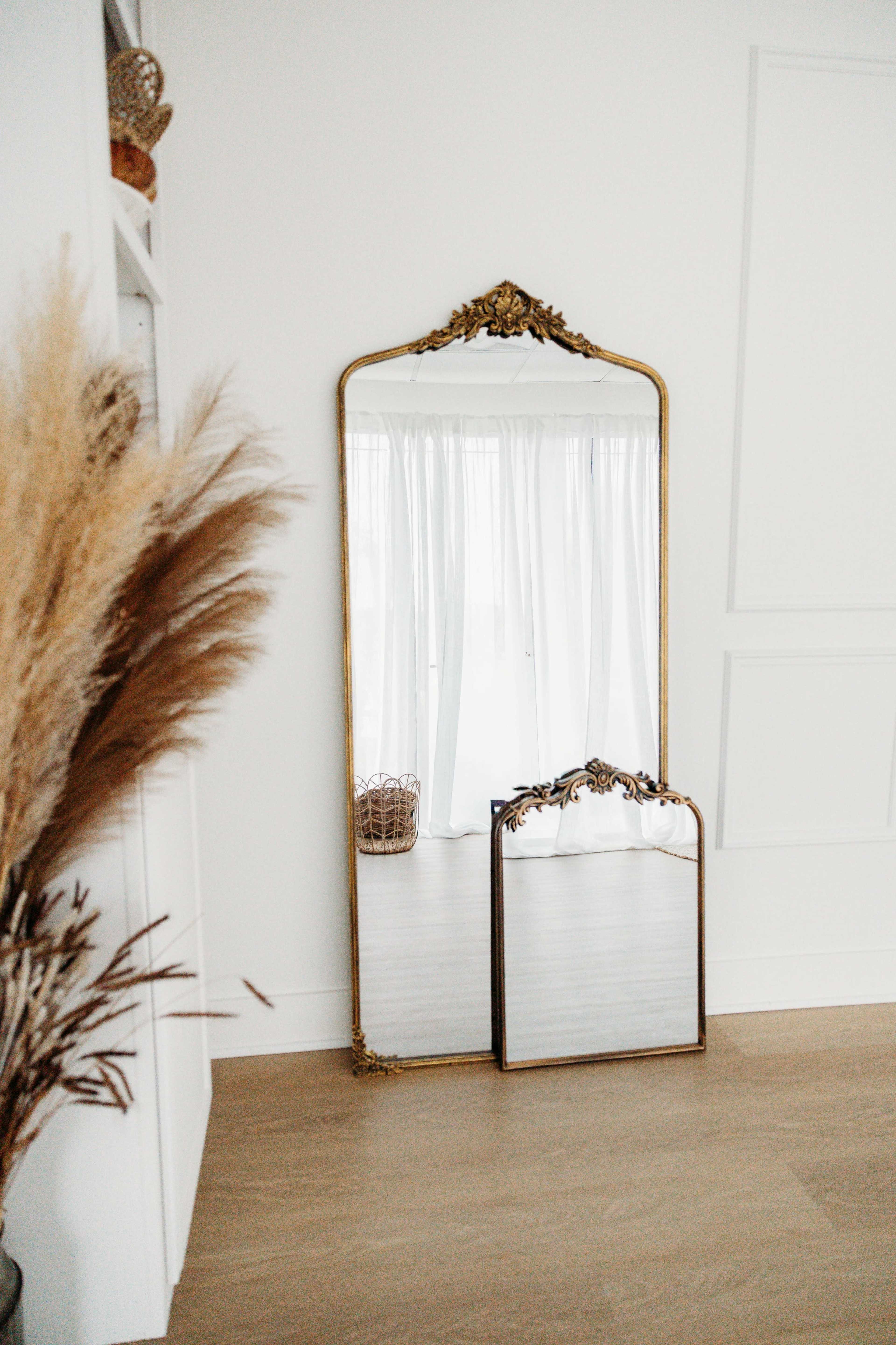 A large ornate mirror stands against a white wall, with a smaller mirror leaning on it, both reflecting light from sheer curtains in the background.