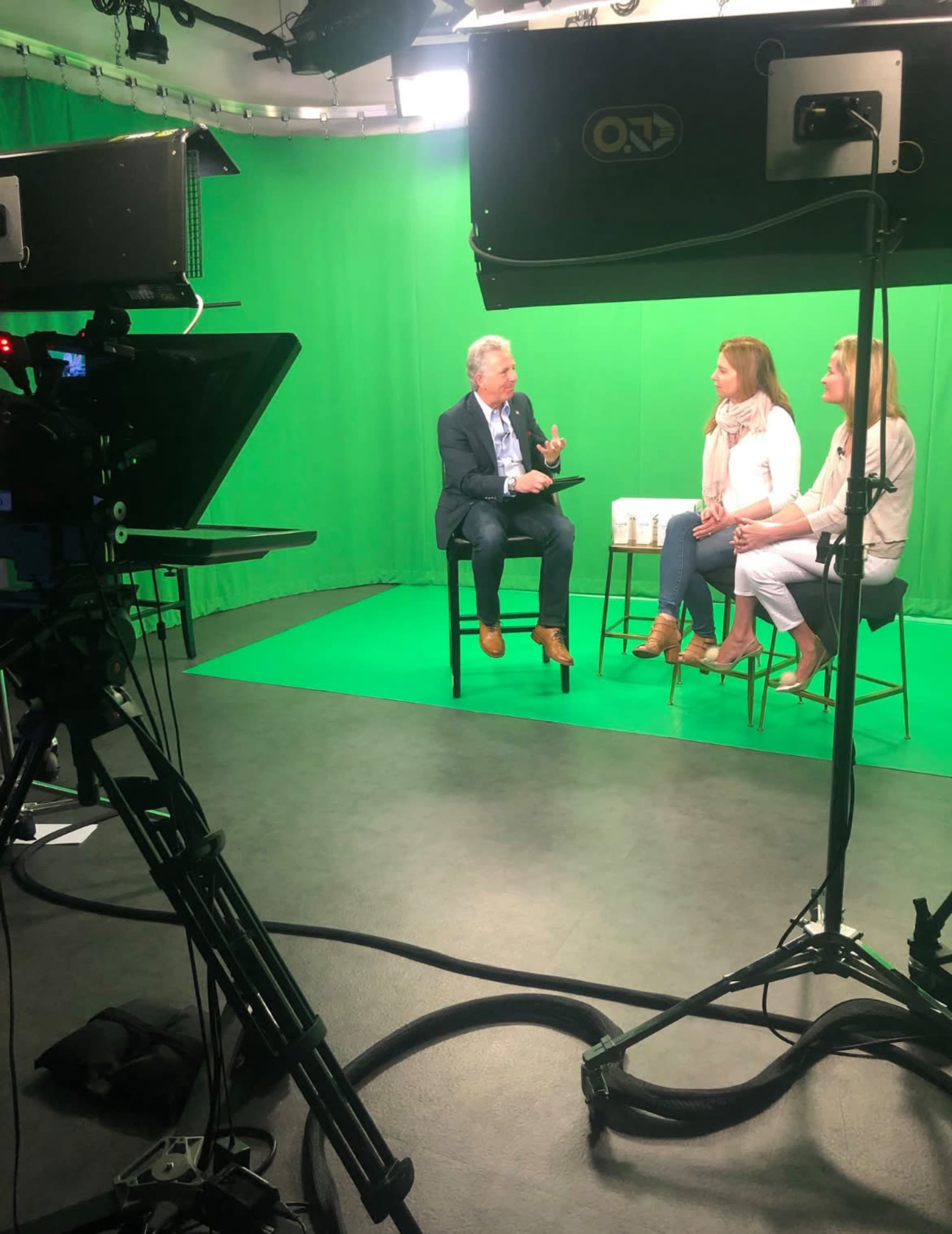 A man and two women sit on high stools in a green-screen studio, engaging in conversation while a camera captures the scene.