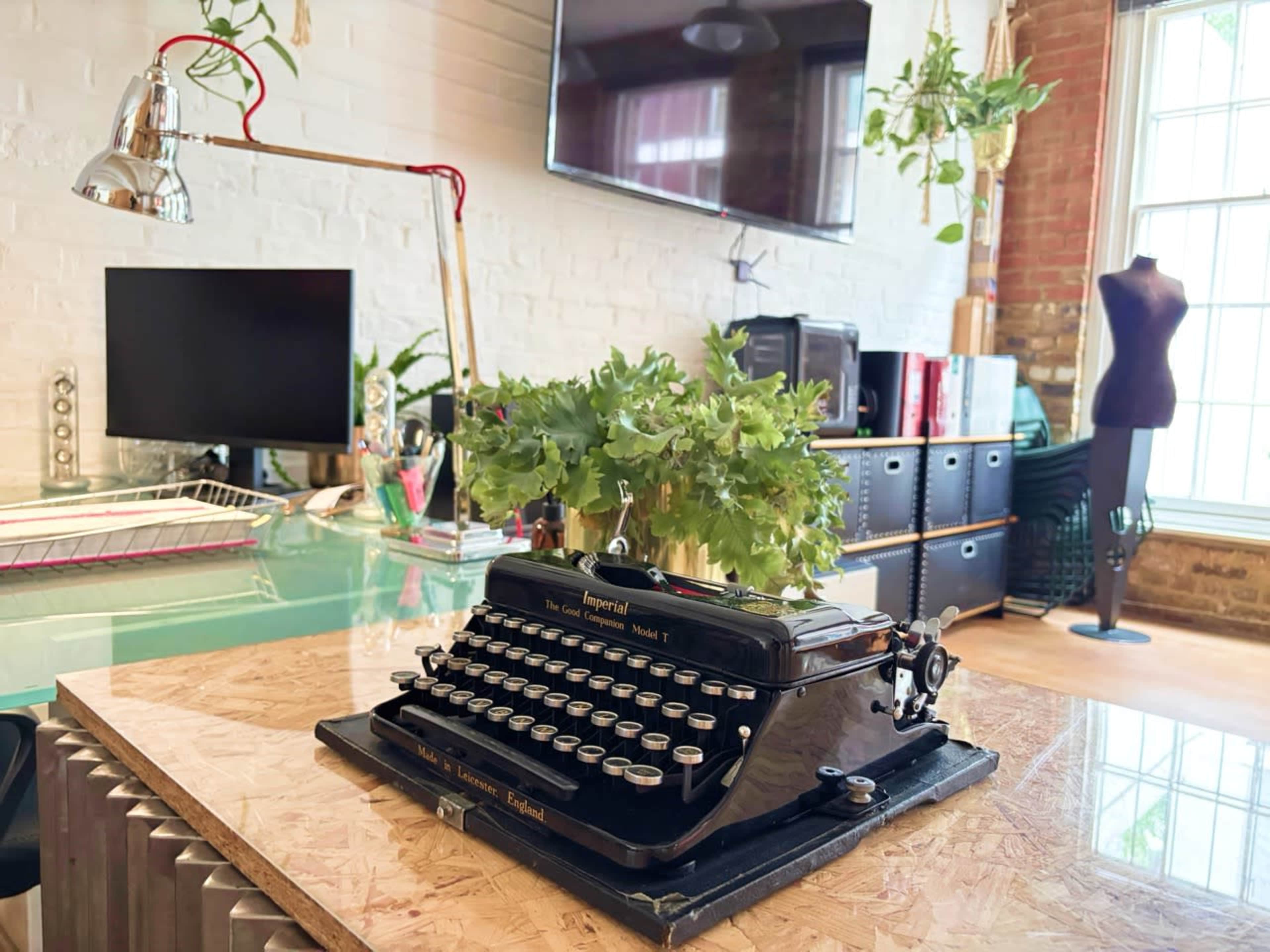 The image shows a vintage typewriter placed on a wooden table surrounded by greenery, with a computer monitor and decorations in a stylish workspace.