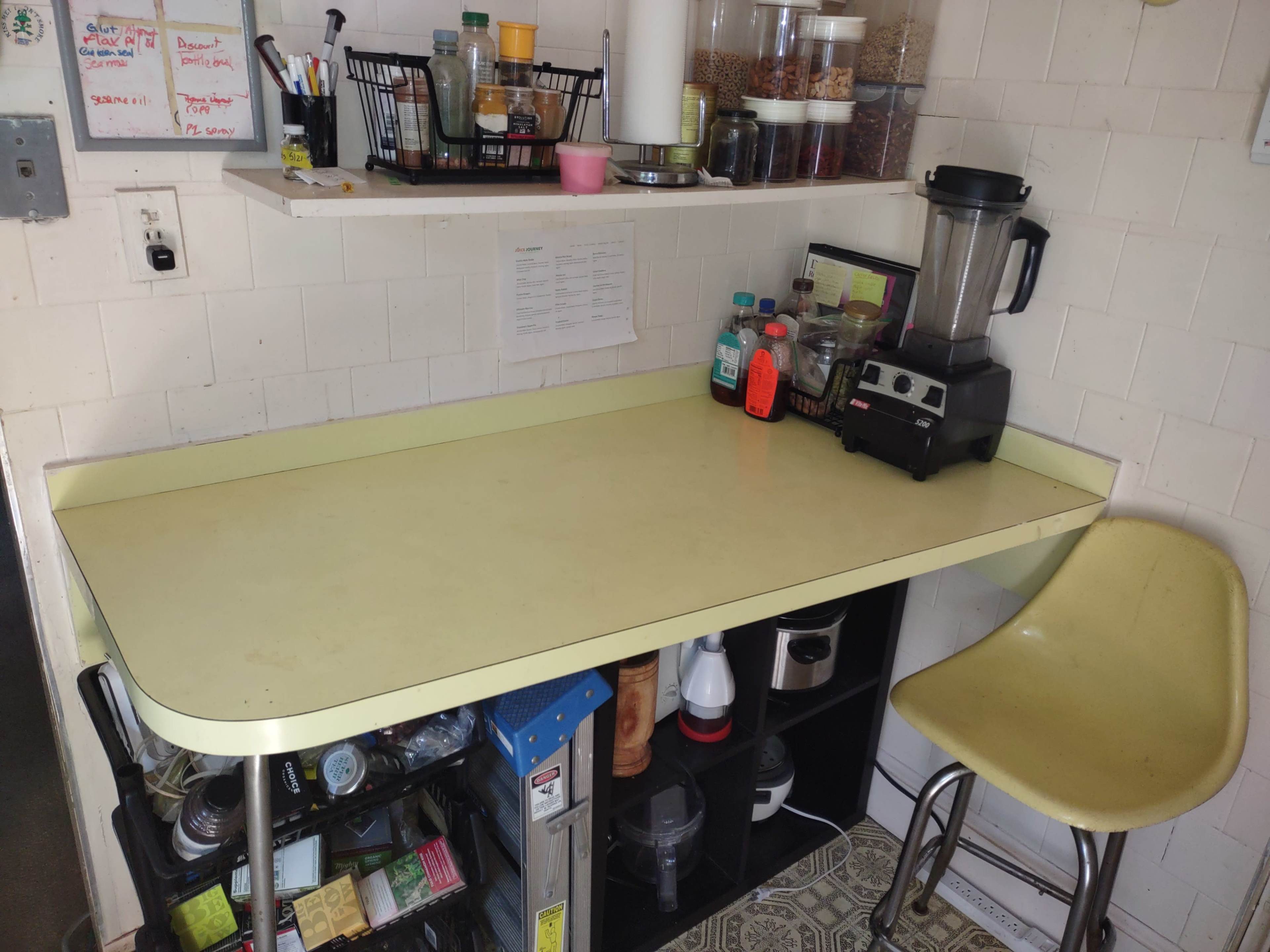A kitchen counter with a yellow surface, a blender, various bottles, and a wall-mounted shelf containing food items and utensils.
