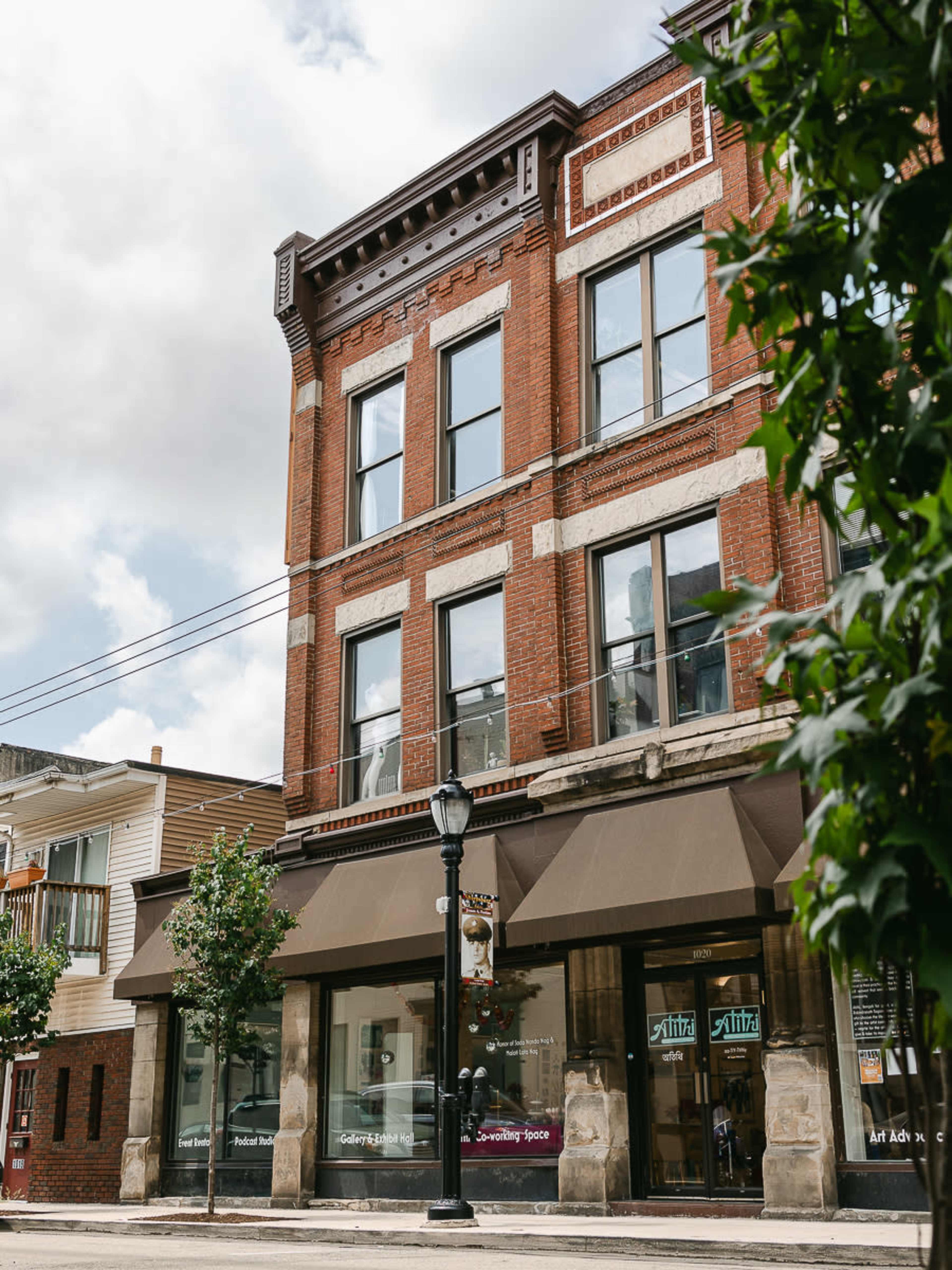The image shows a historic brick building with large windows and a decorative facade, located on a street corner with shops on the ground floor.
