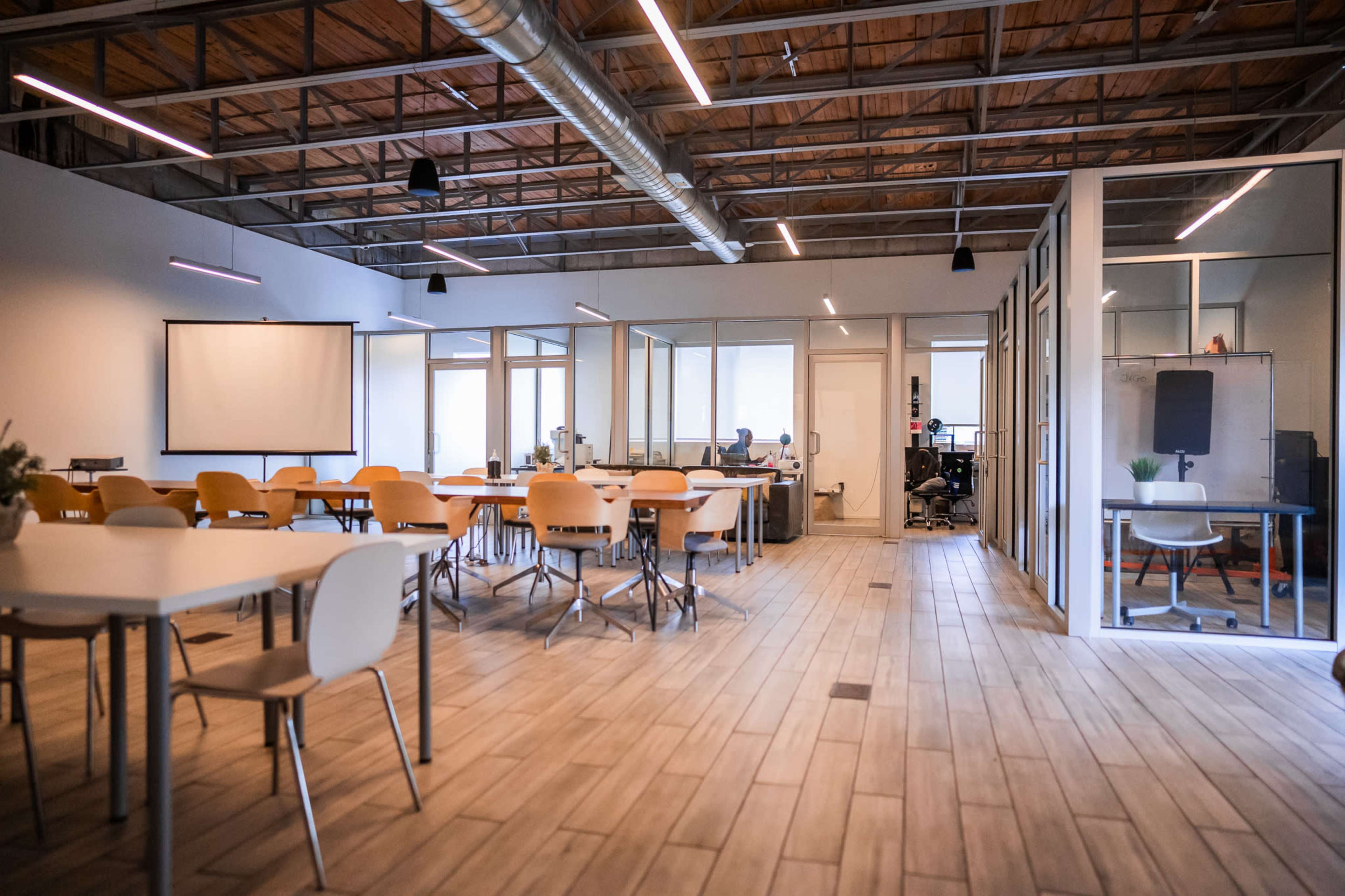 A spacious office interior features rows of tables and chairs, with a glass-walled meeting room and workstations visible in the background.