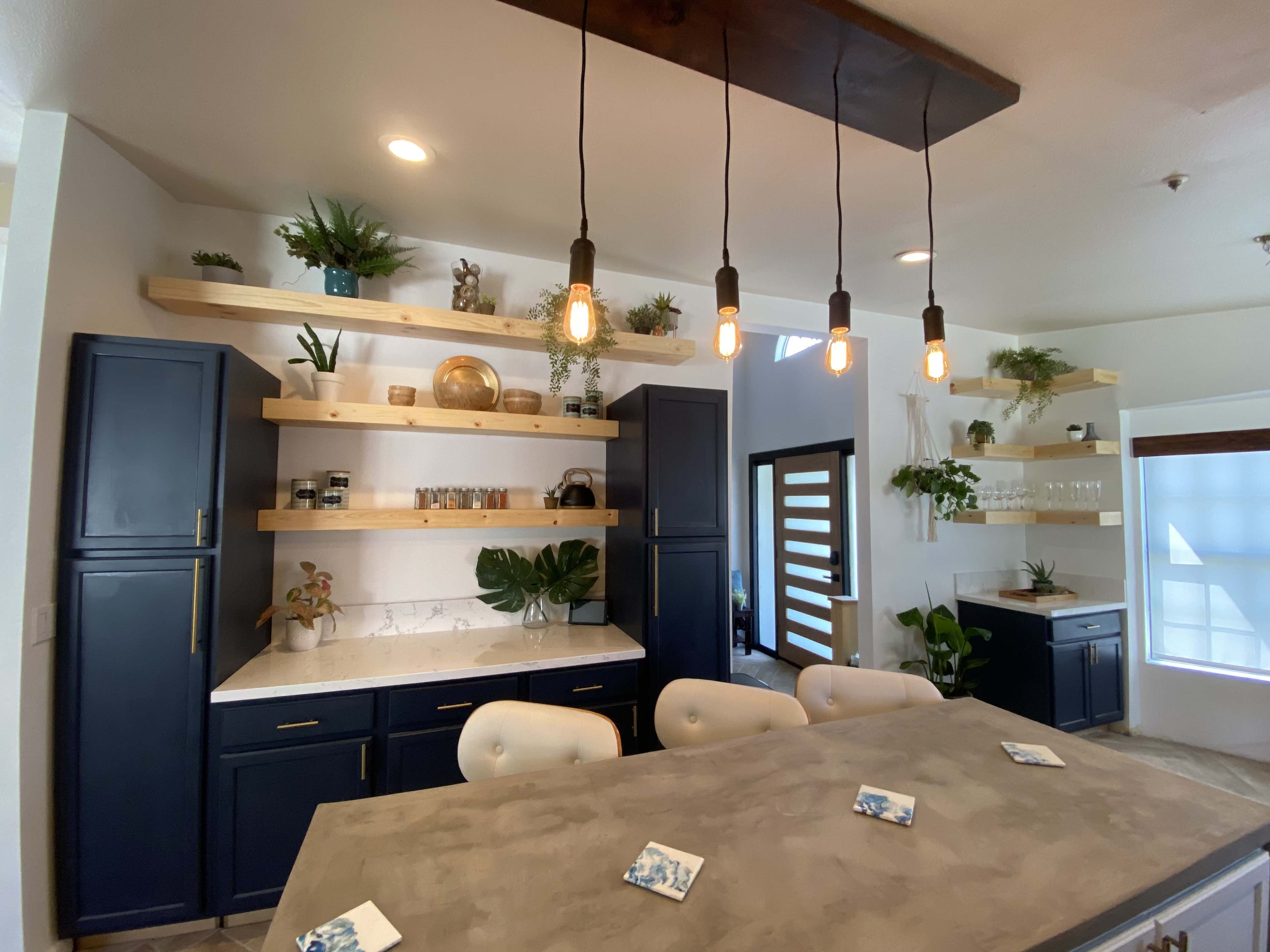 A modern kitchen with blue cabinetry, a centerpiece island, wooden shelves adorned with plants and decorative items, and pendant lighting.