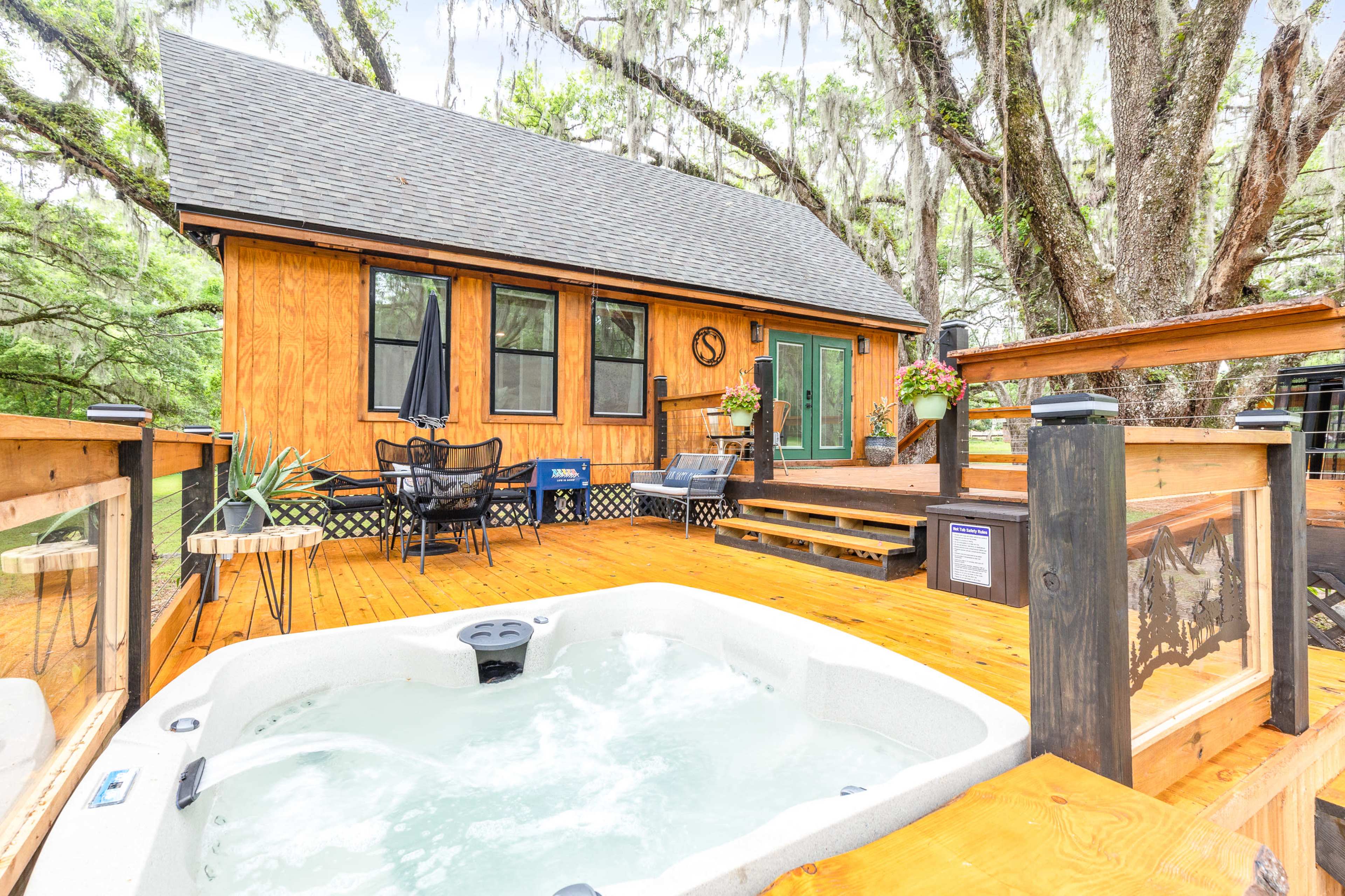 A wooden deck with a hot tub is situated in front of a cabin, surrounded by trees with hanging moss.