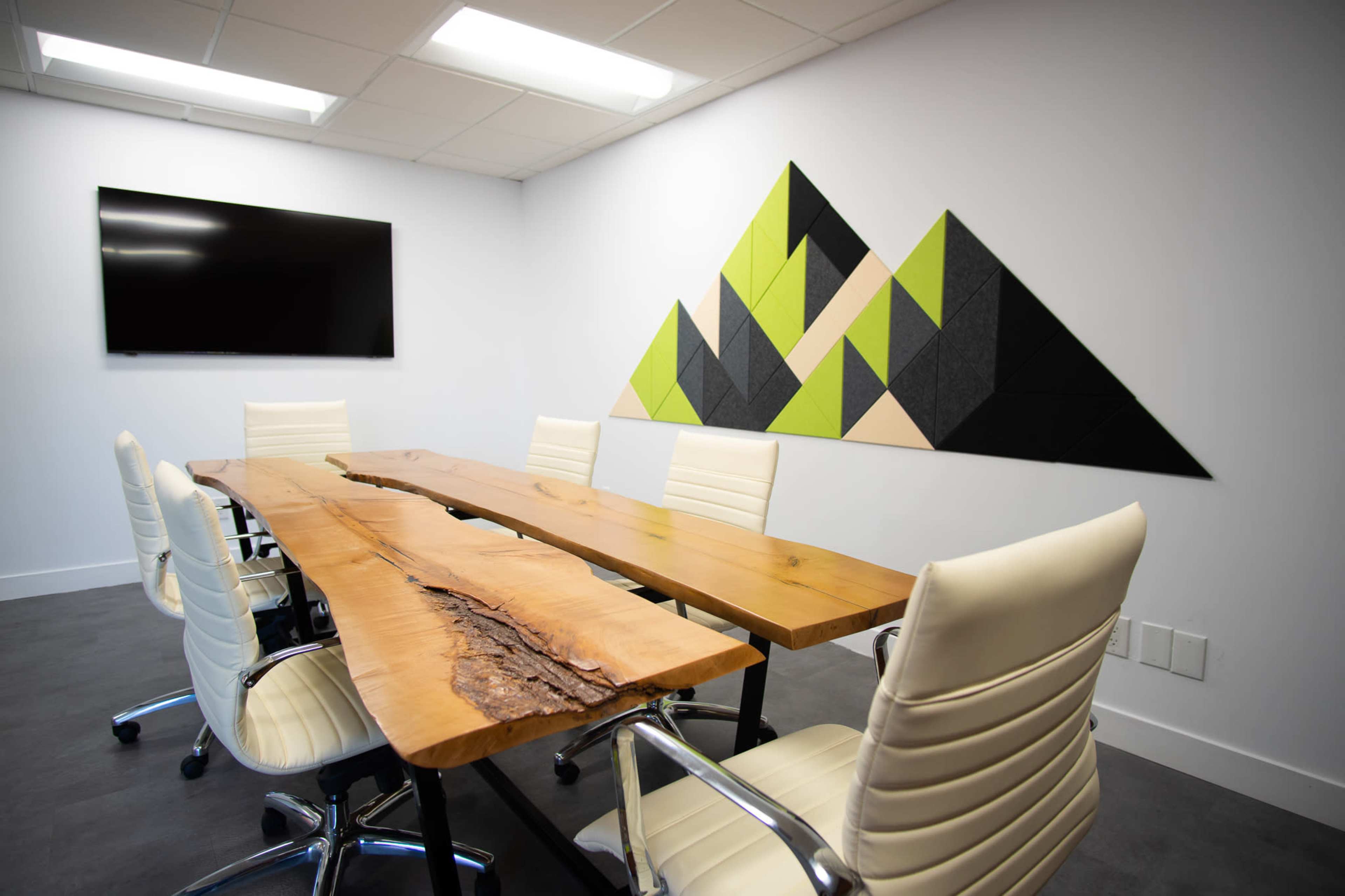 A modern conference room features a long wooden table with six white chairs and a wall decorated with triangular felt panels in green and black, alongside a mounted television.