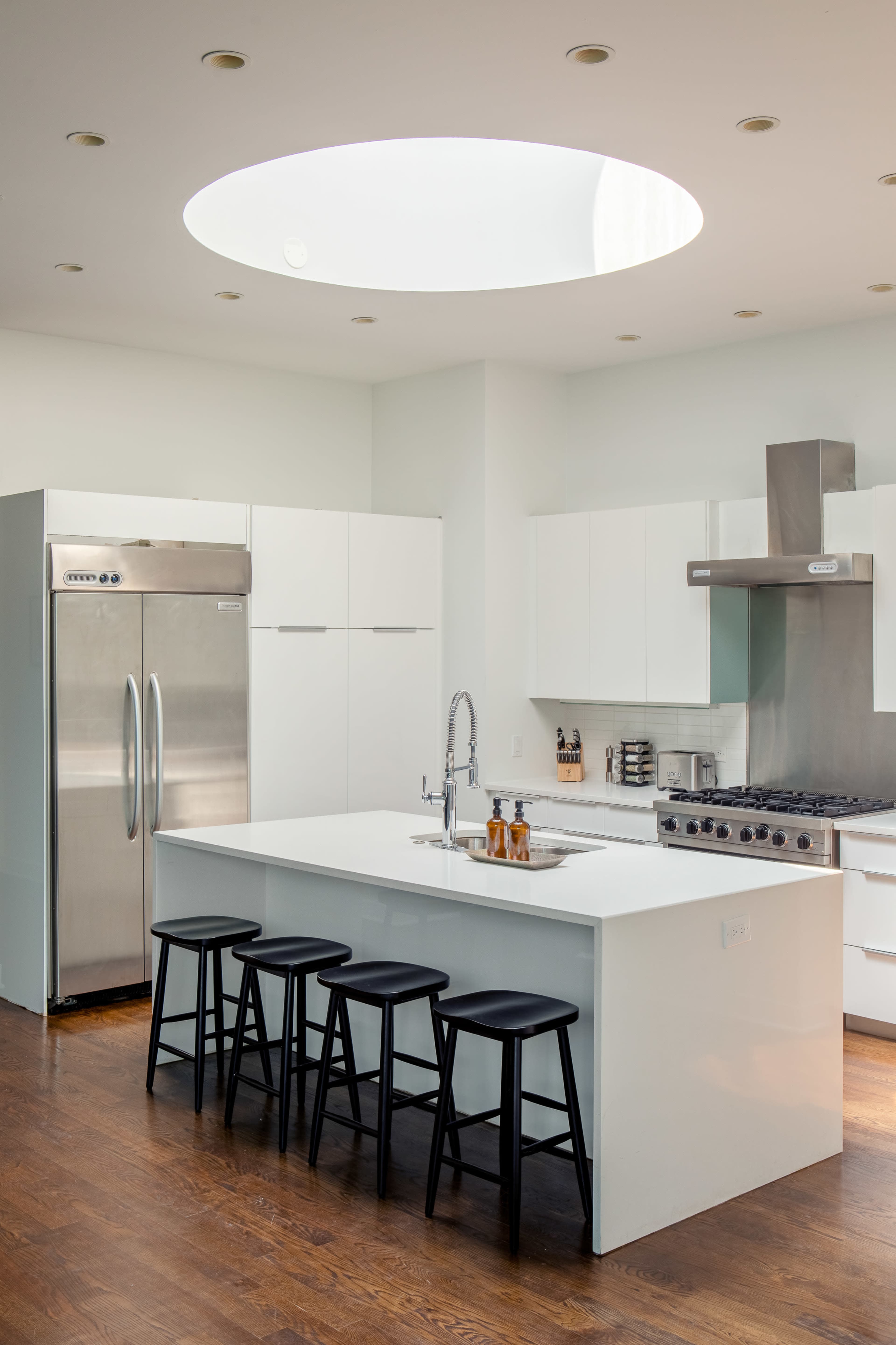 A modern kitchen features white cabinetry, stainless steel appliances, and a central island with black stools, illuminated by a circular skylight.
