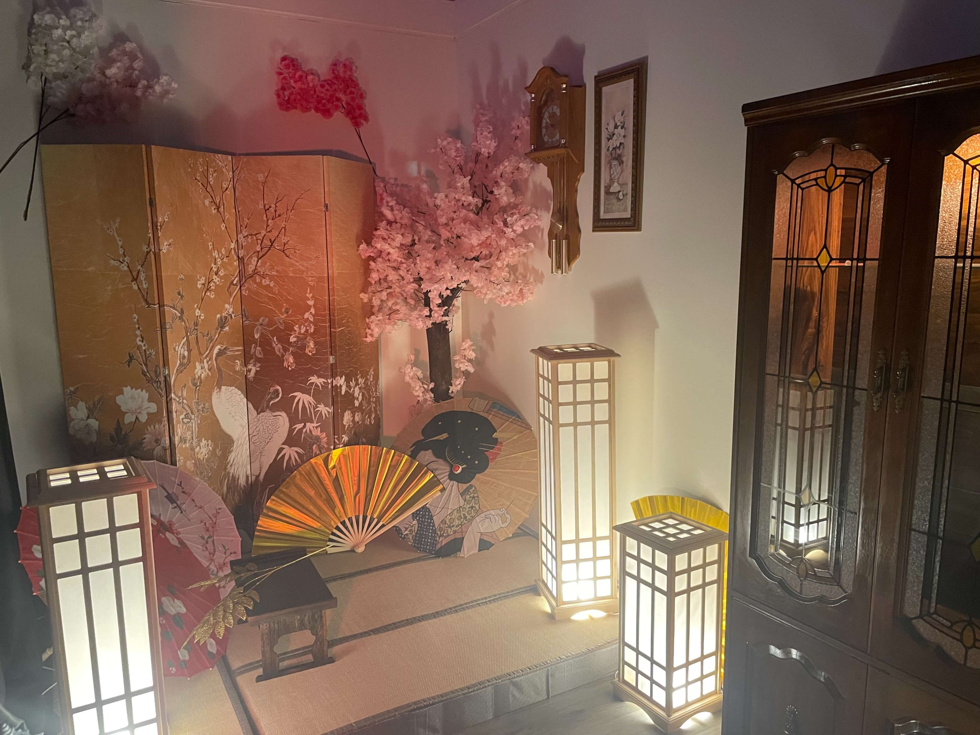 The scene features a cozy room adorned with a folding screen, decorative fans, cherry blossom branches, lanterns, and a wooden cabinet.