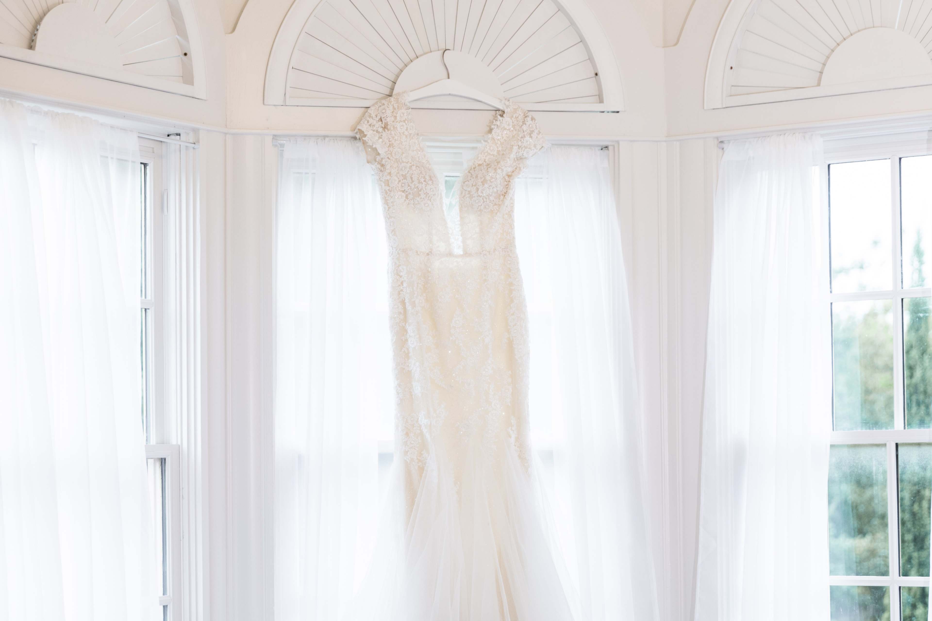 A bridal gown hangs on a hanger against a backdrop of flowing white curtains.
