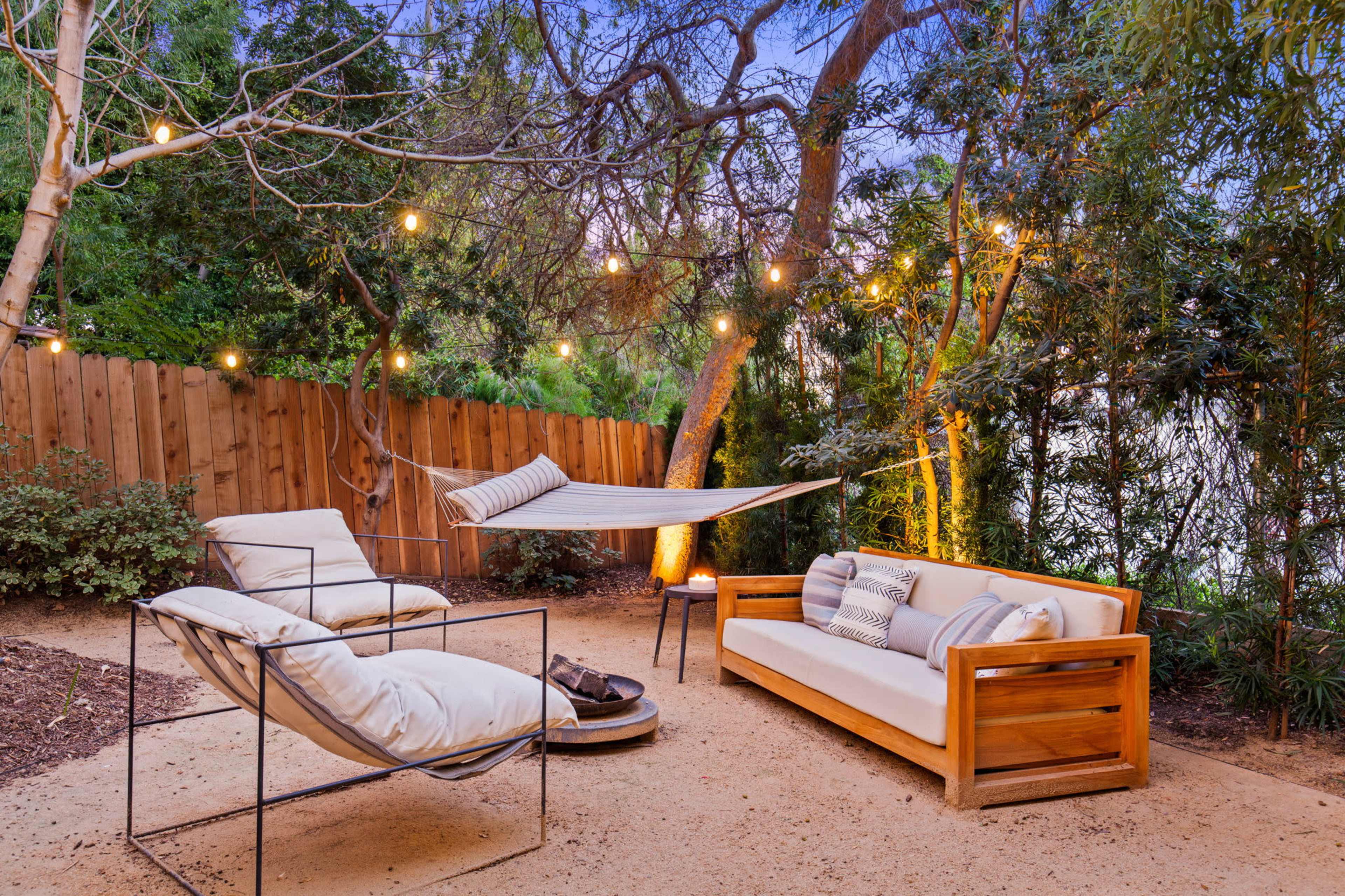 A cozy outdoor space features a hammock, two armchairs, and a sofa surrounded by string lights and lush greenery.
