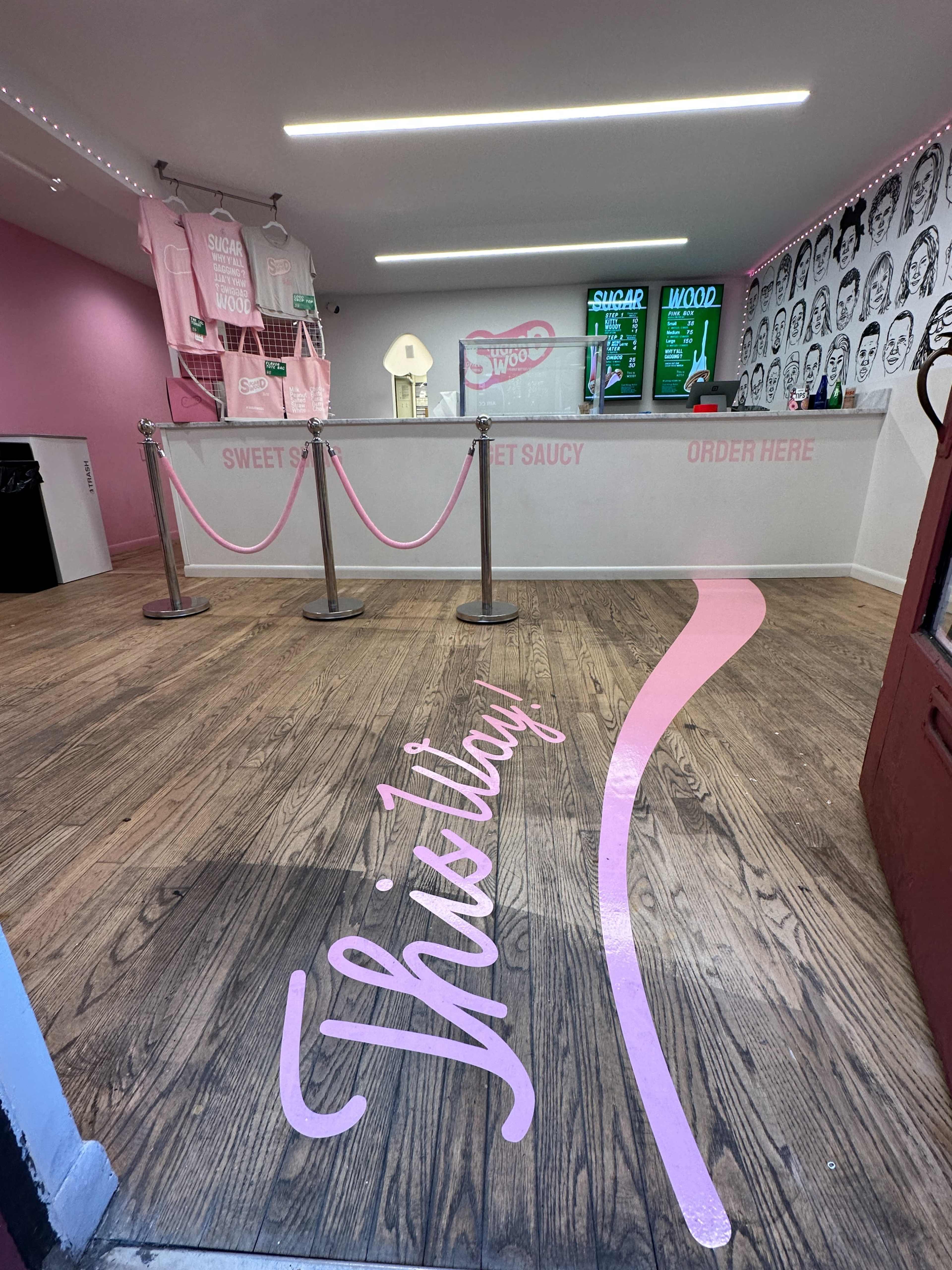 The interior of a brightly colored dessert shop with a wooden floor, featuring a counter with menus and a pink pathway leading toward it.