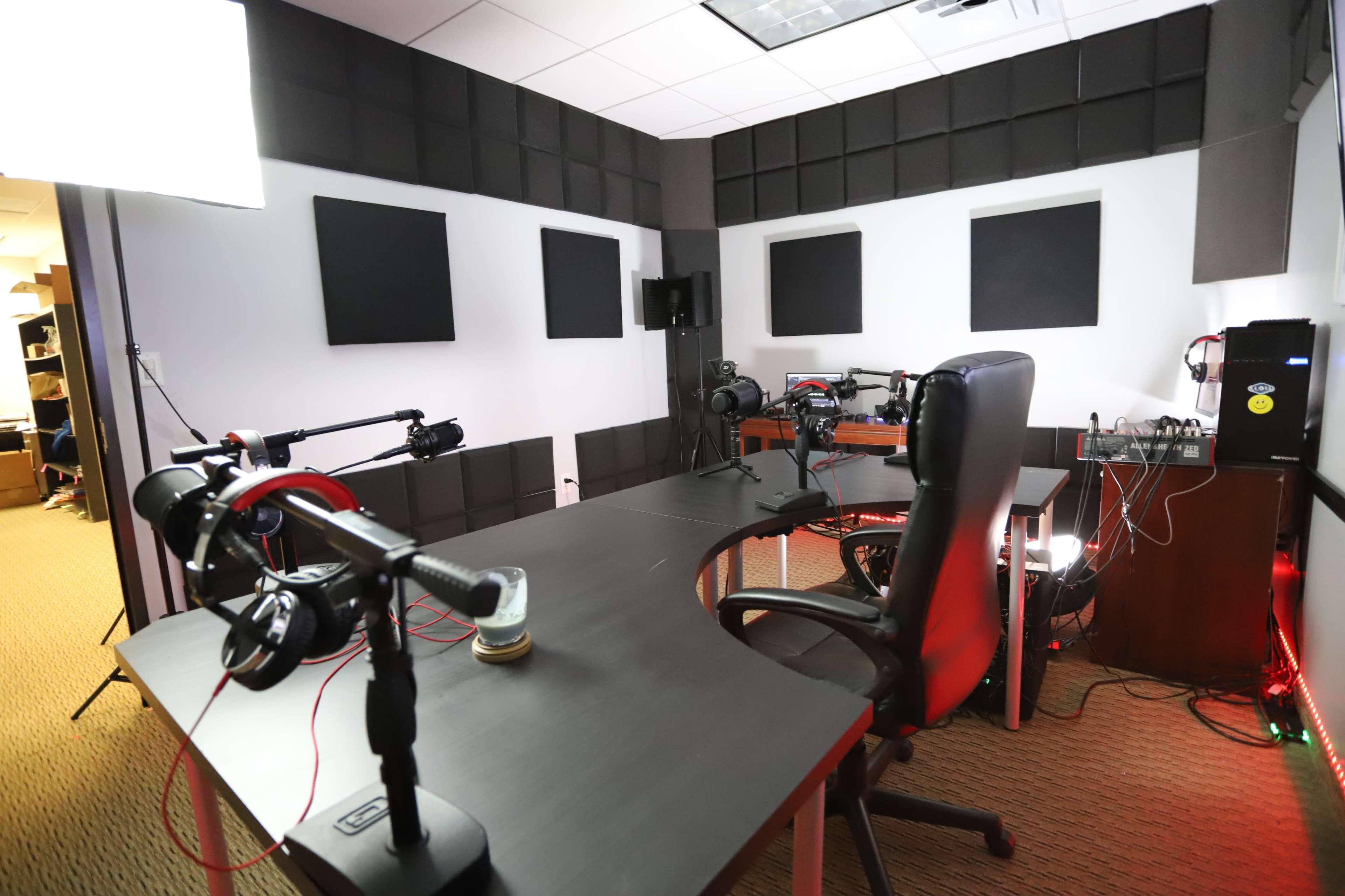 The image shows a recording studio featuring black soundproof panels on the walls, a large desk with multiple microphones and headphones, and a central black office chair.