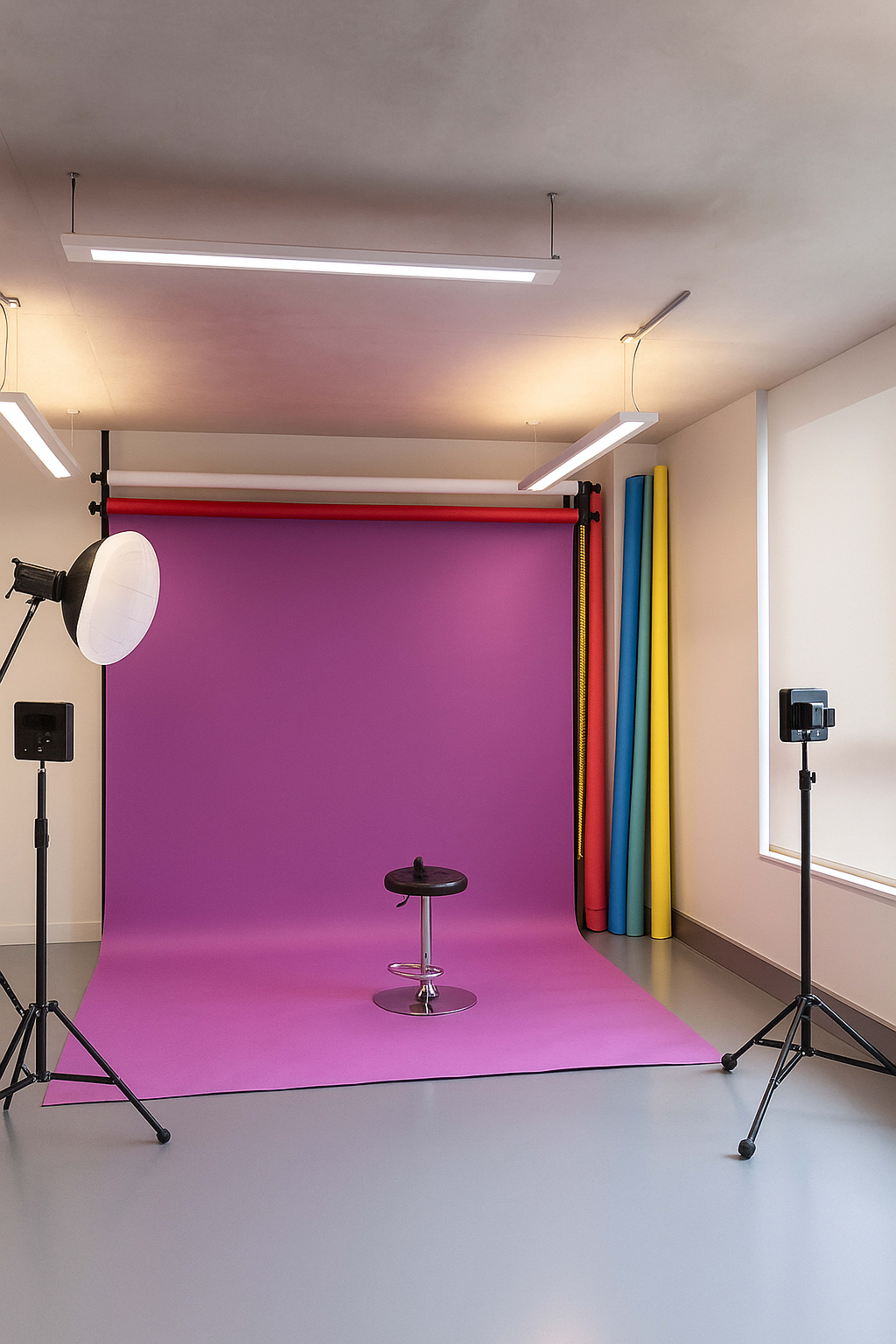 A photography studio features a purple backdrop, a black stool, and colored roll backdrops with lighting equipment positioned around the space.