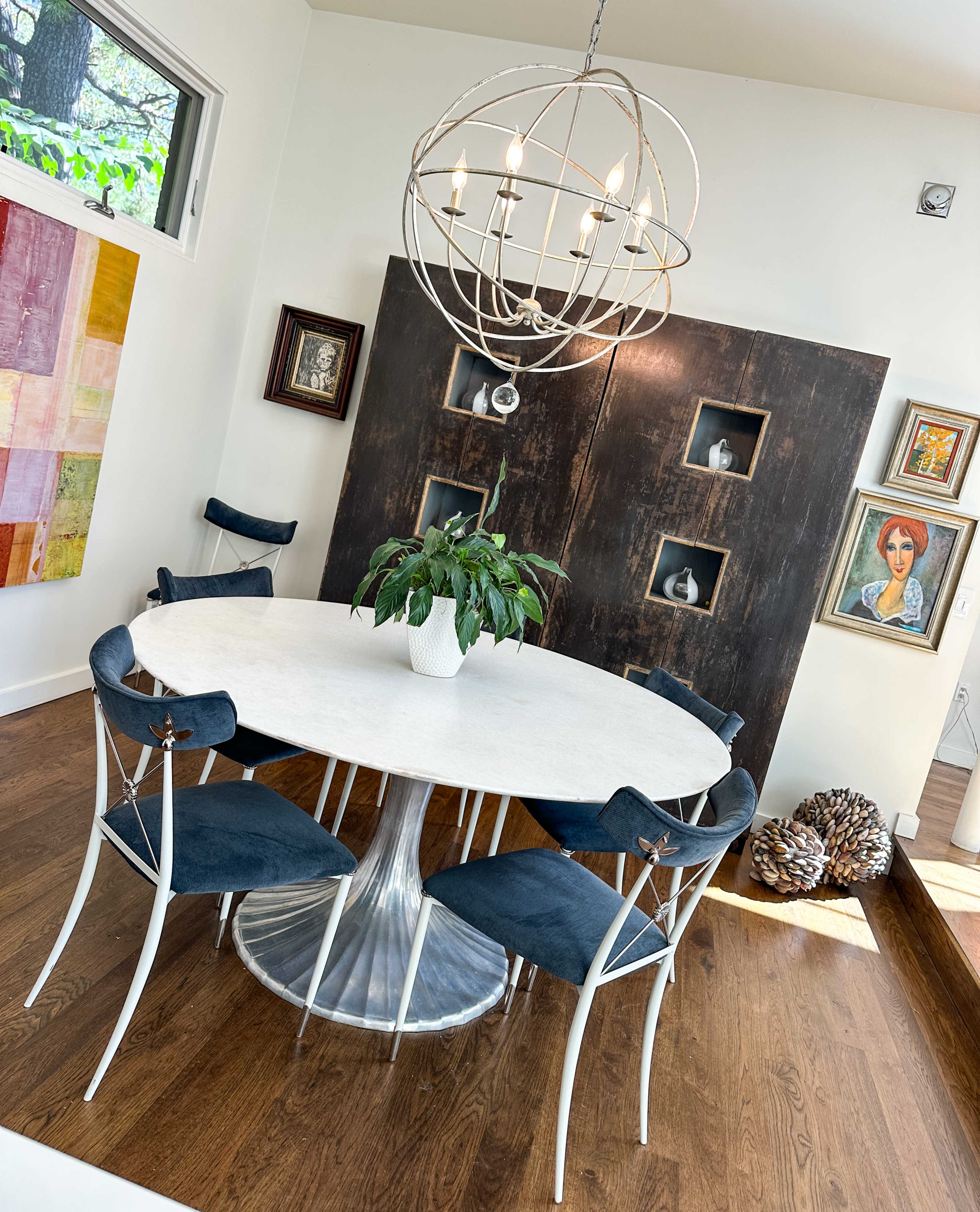 A round white table with dark blue chairs is set in a bright room featuring a large spherical light fixture and a wooden panel display with framed artworks.