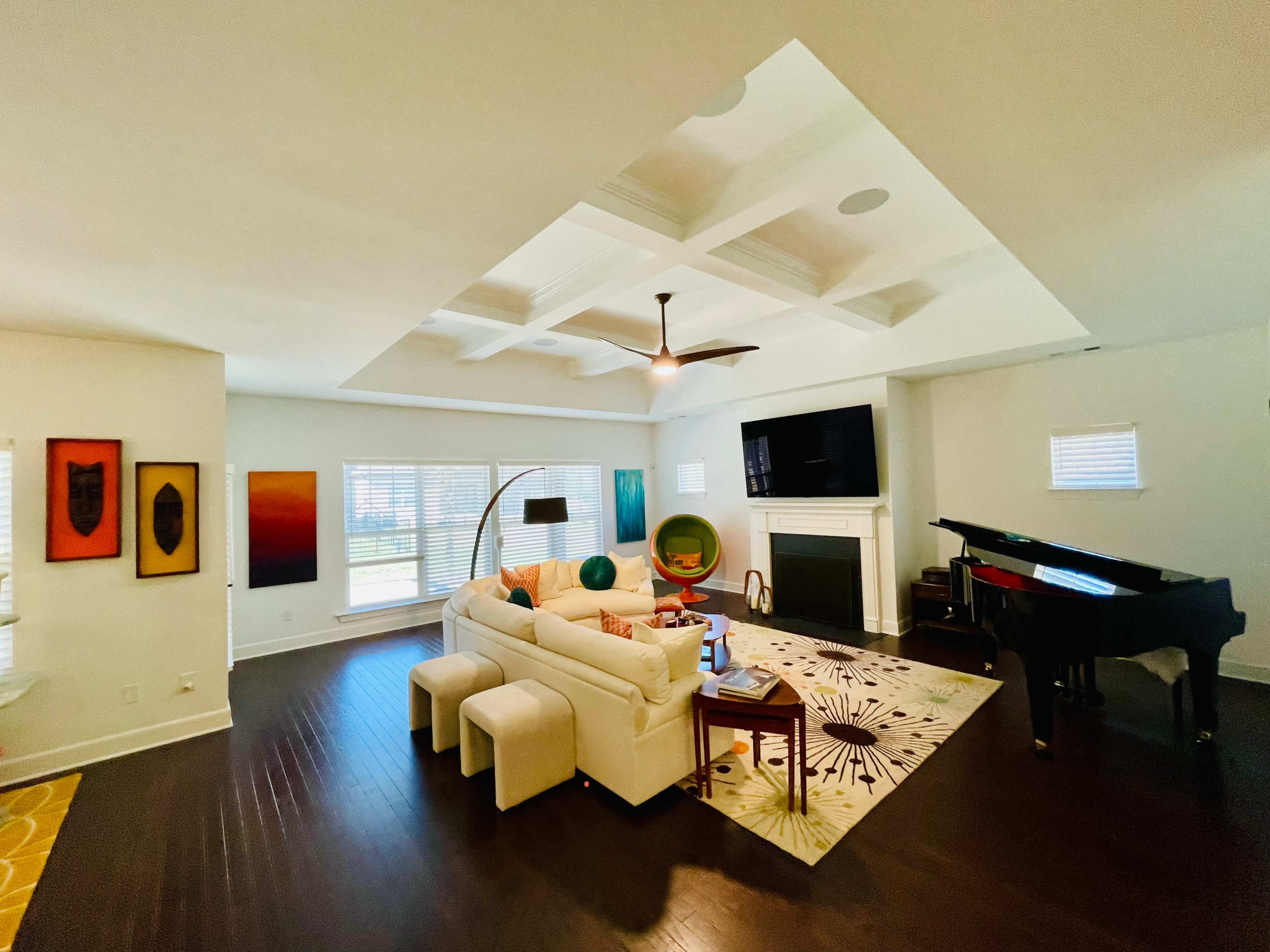 A bright living room features a white sectional sofa, a grand piano, and large windows with a view of the outdoors.
