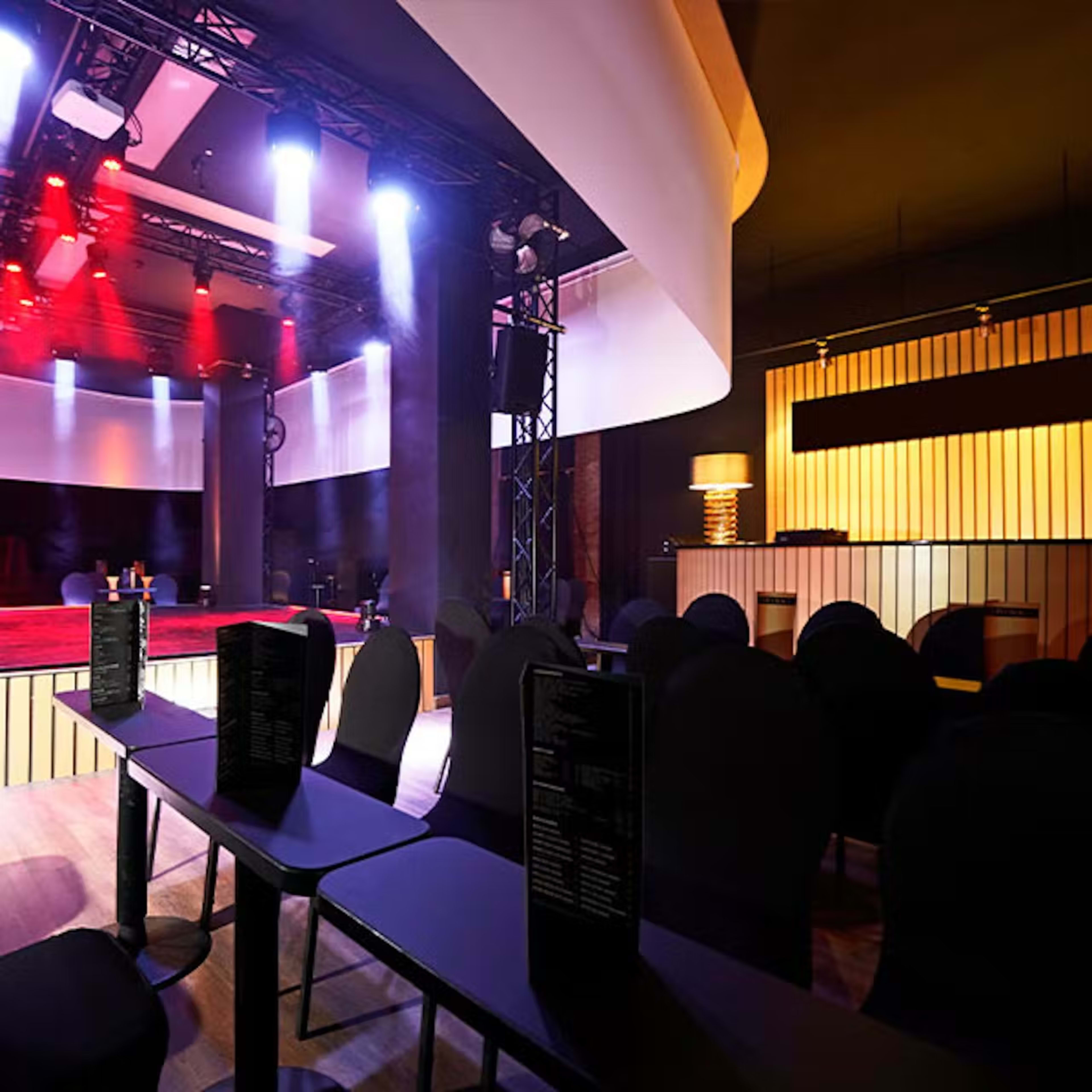 The image shows an empty performance space with rows of black chairs and tables, illuminated by stage lights and featuring a bar area in the background.