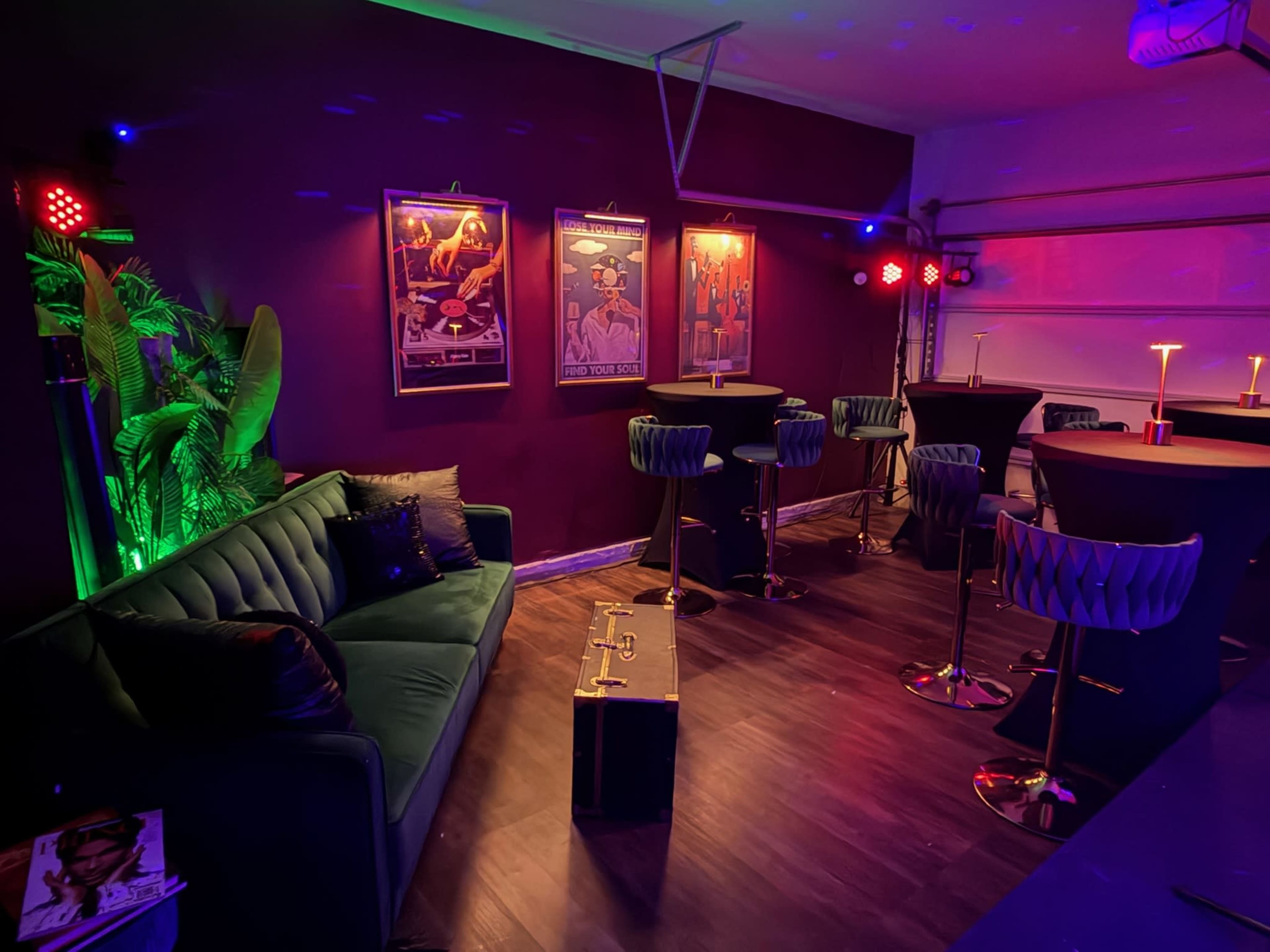 The image shows a stylish lounge area with a dark purple wall, modern bar stools, a green couch, and illuminated posters.