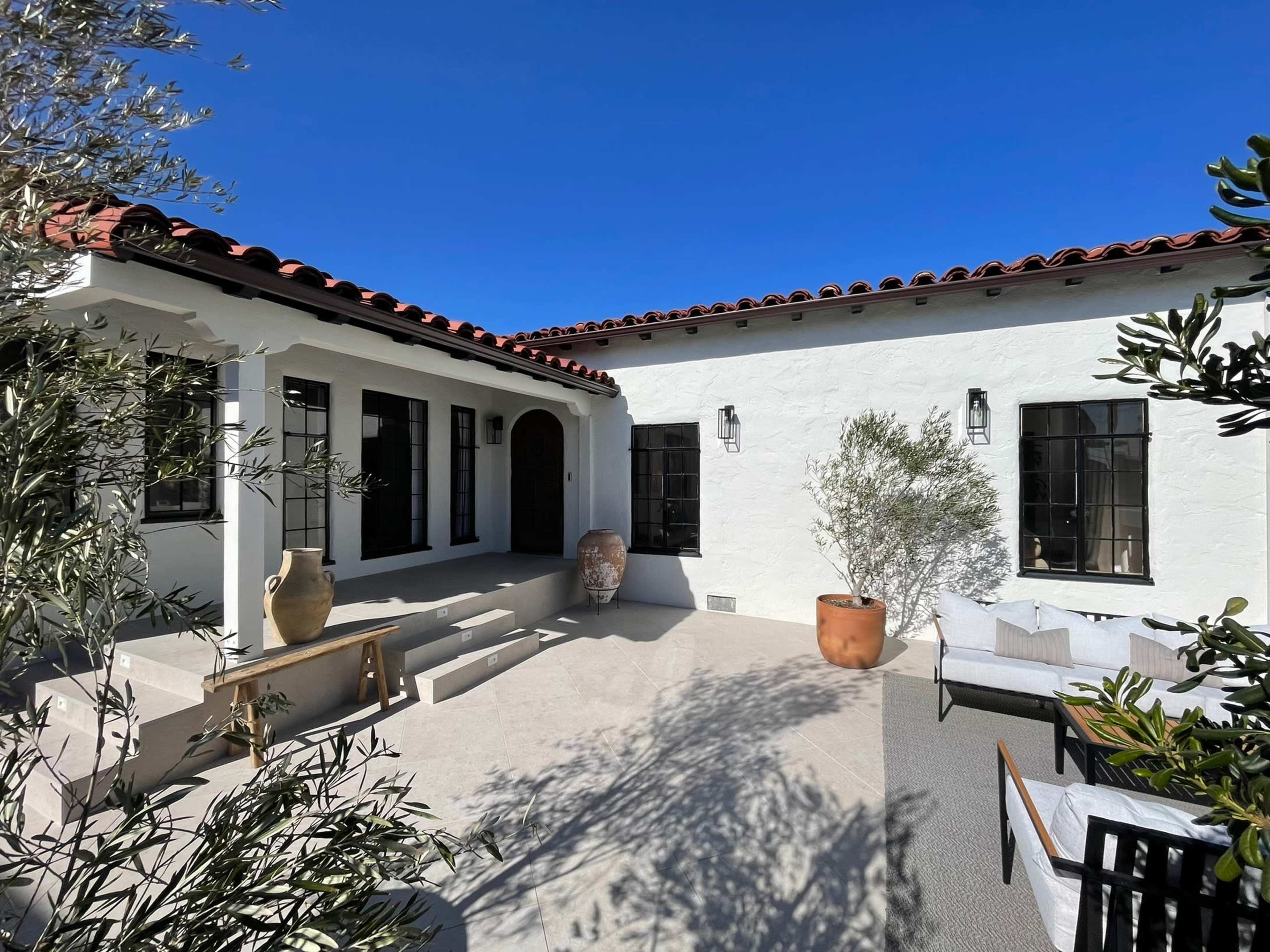 A sunlit courtyard features a white adobe-style house with a red tile roof, surrounded by potted plants and outdoor seating.