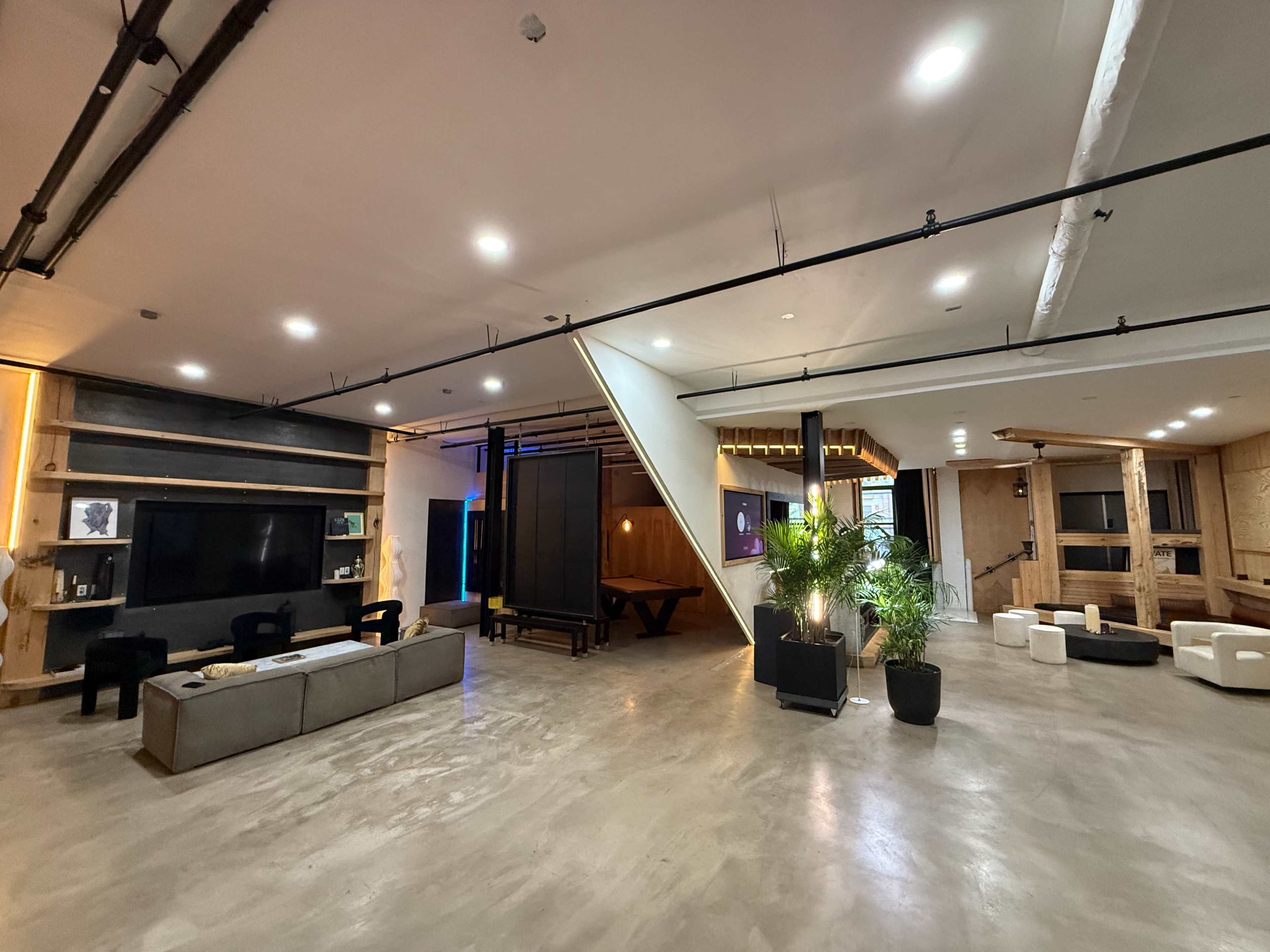 The image shows a modern, open-concept space with a blend of wood and concrete elements, featuring seating areas, a large television, and a staircase.