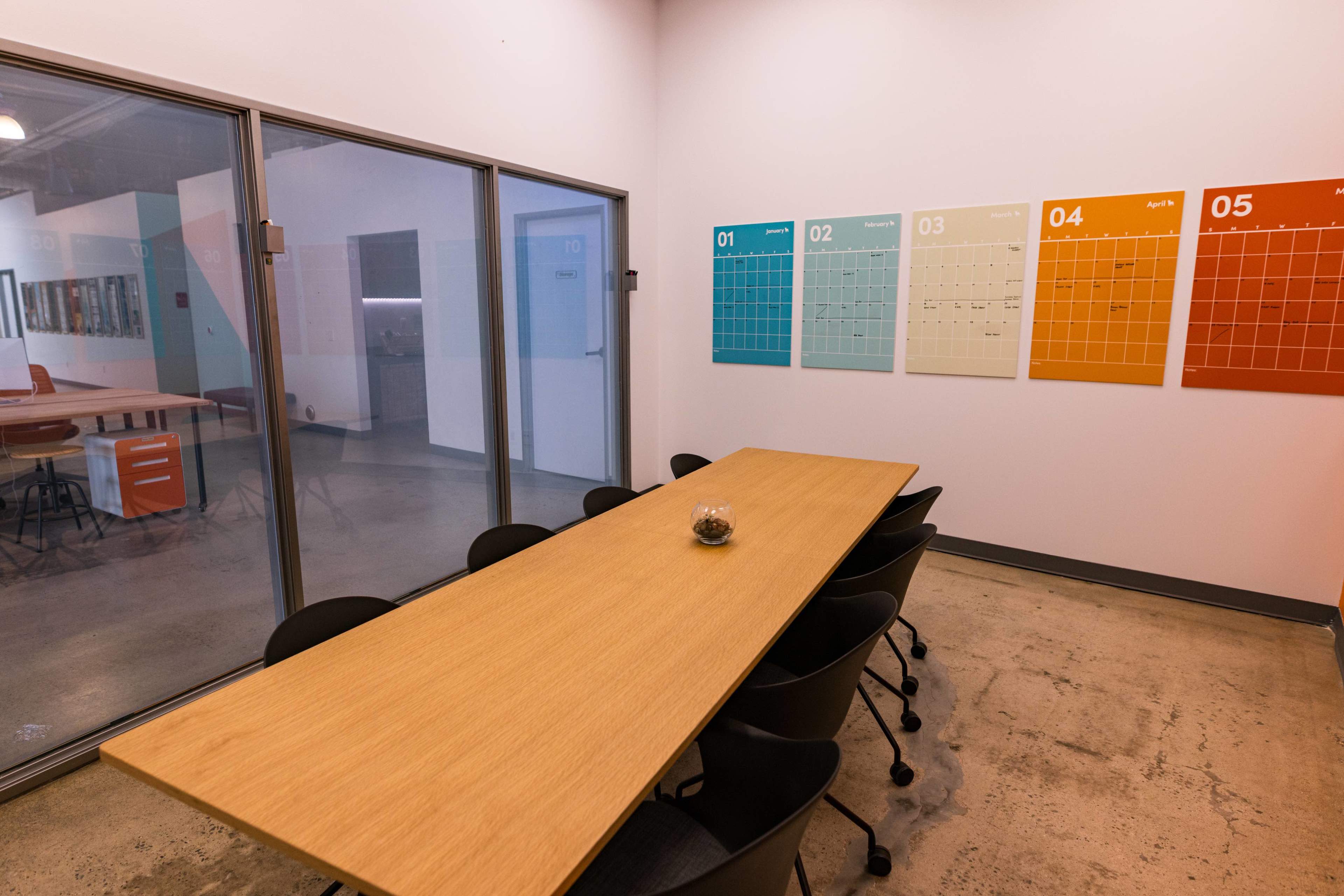 A conference room features a long wooden table surrounded by black chairs, with colorful monthly calendars mounted on the wall.