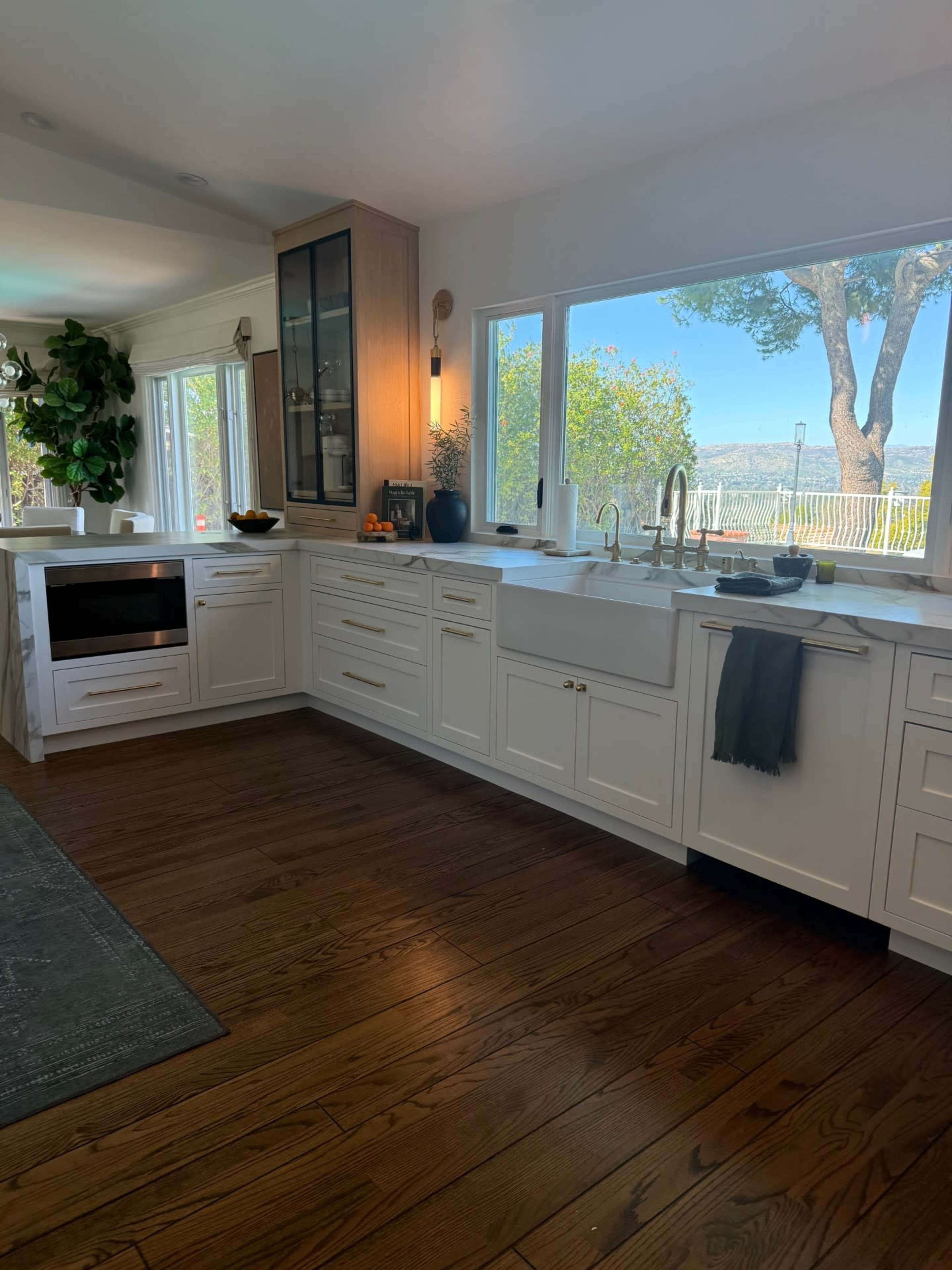 The kitchen features white cabinetry, a large window overlooking a scenic view, and wooden flooring.