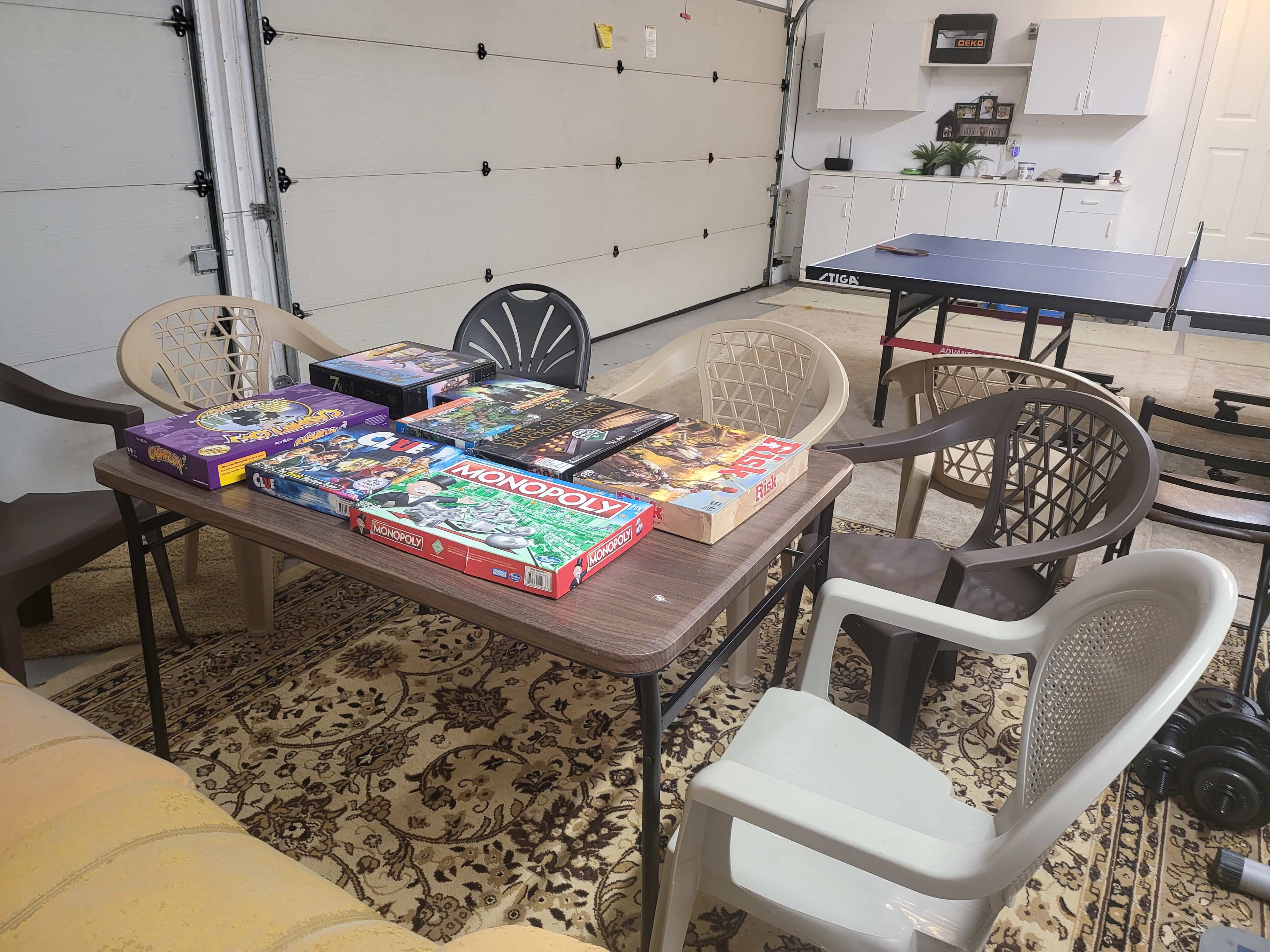 A table with several board games is surrounded by plastic chairs in a spacious garage.