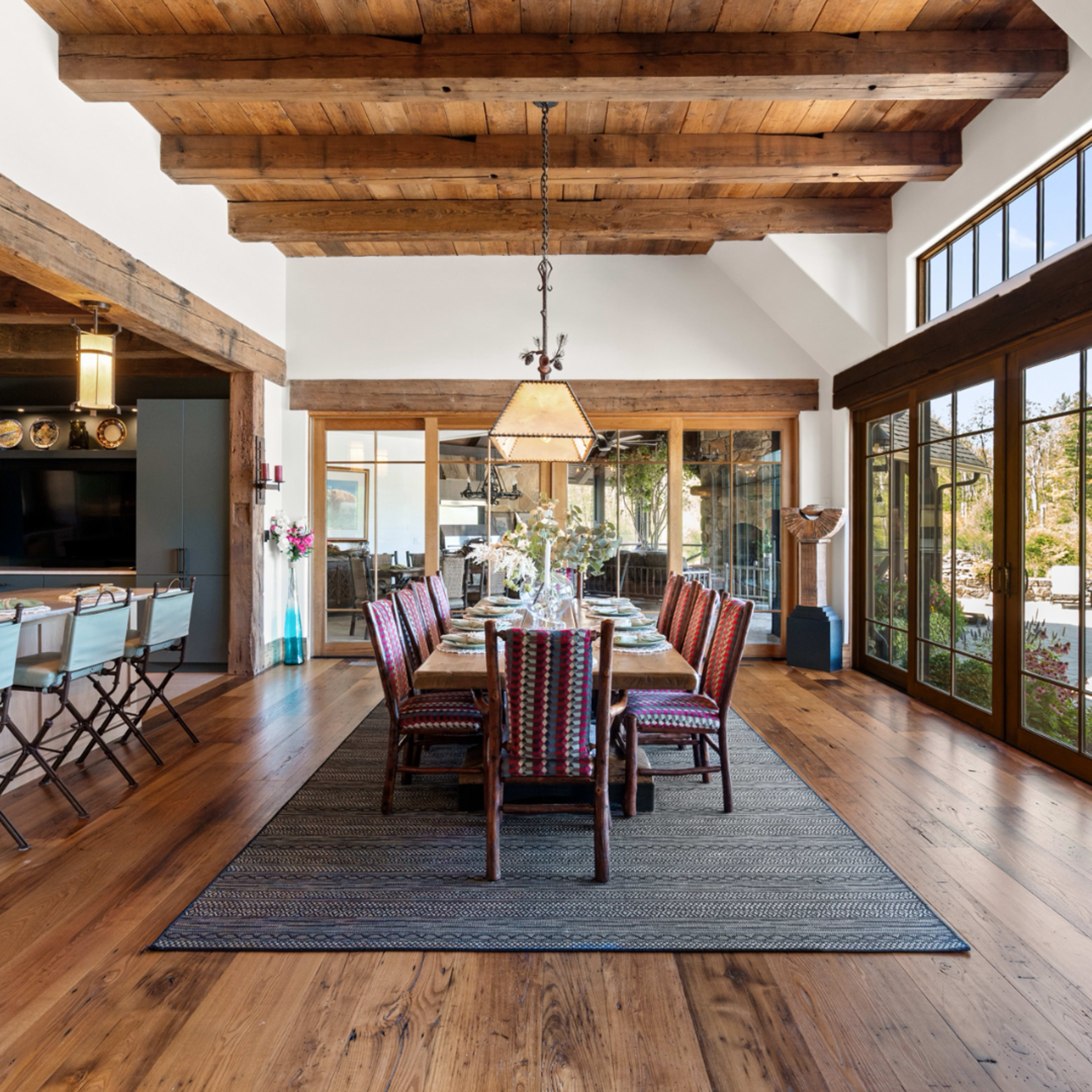 The image shows a spacious dining area with a long wooden table surrounded by colorful chairs, tall windows, and exposed wooden beams on the ceiling.