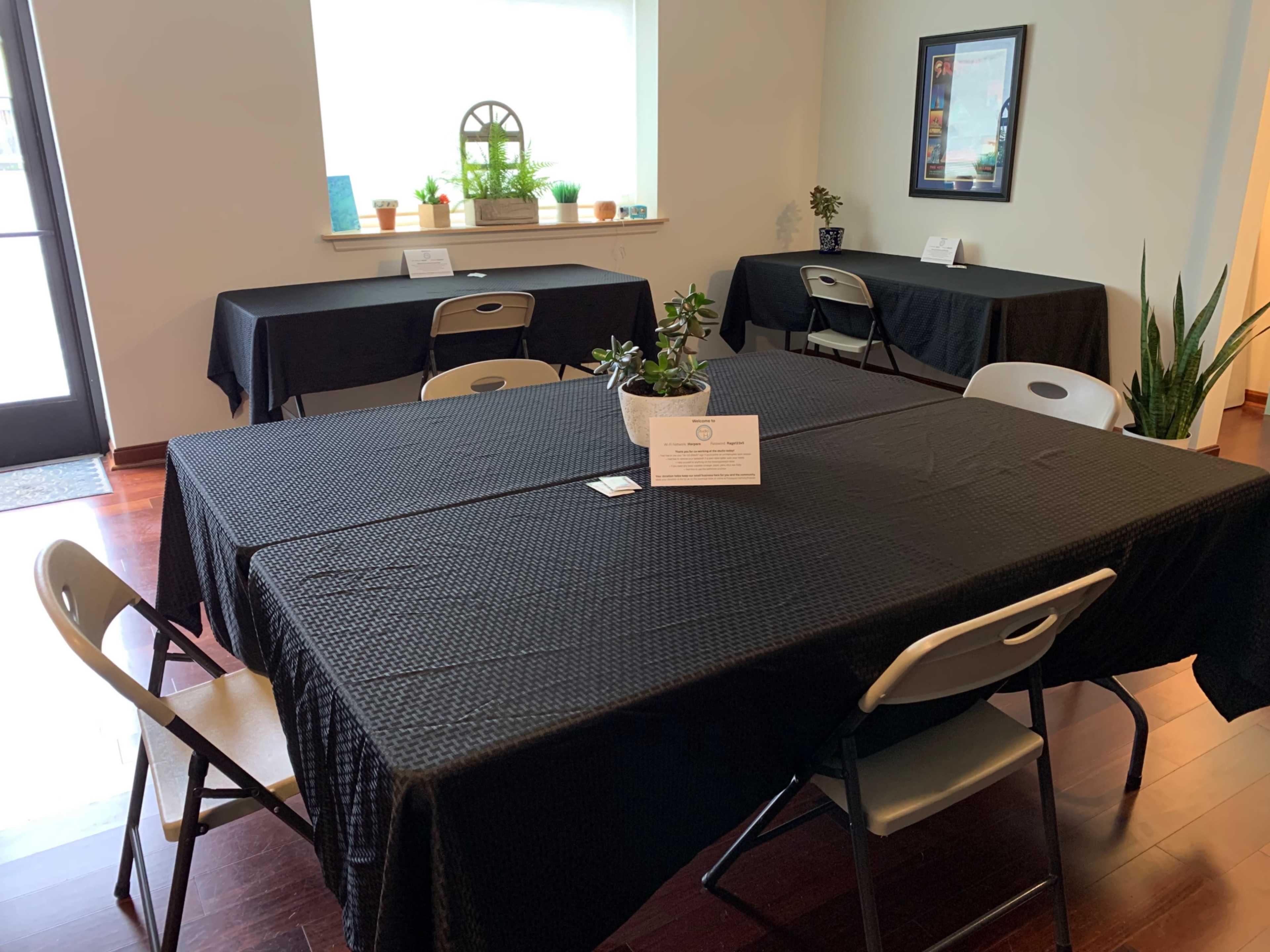 The image shows a spacious room with several tables covered in black tablecloths, arranged with chairs around them, and plants near a window.