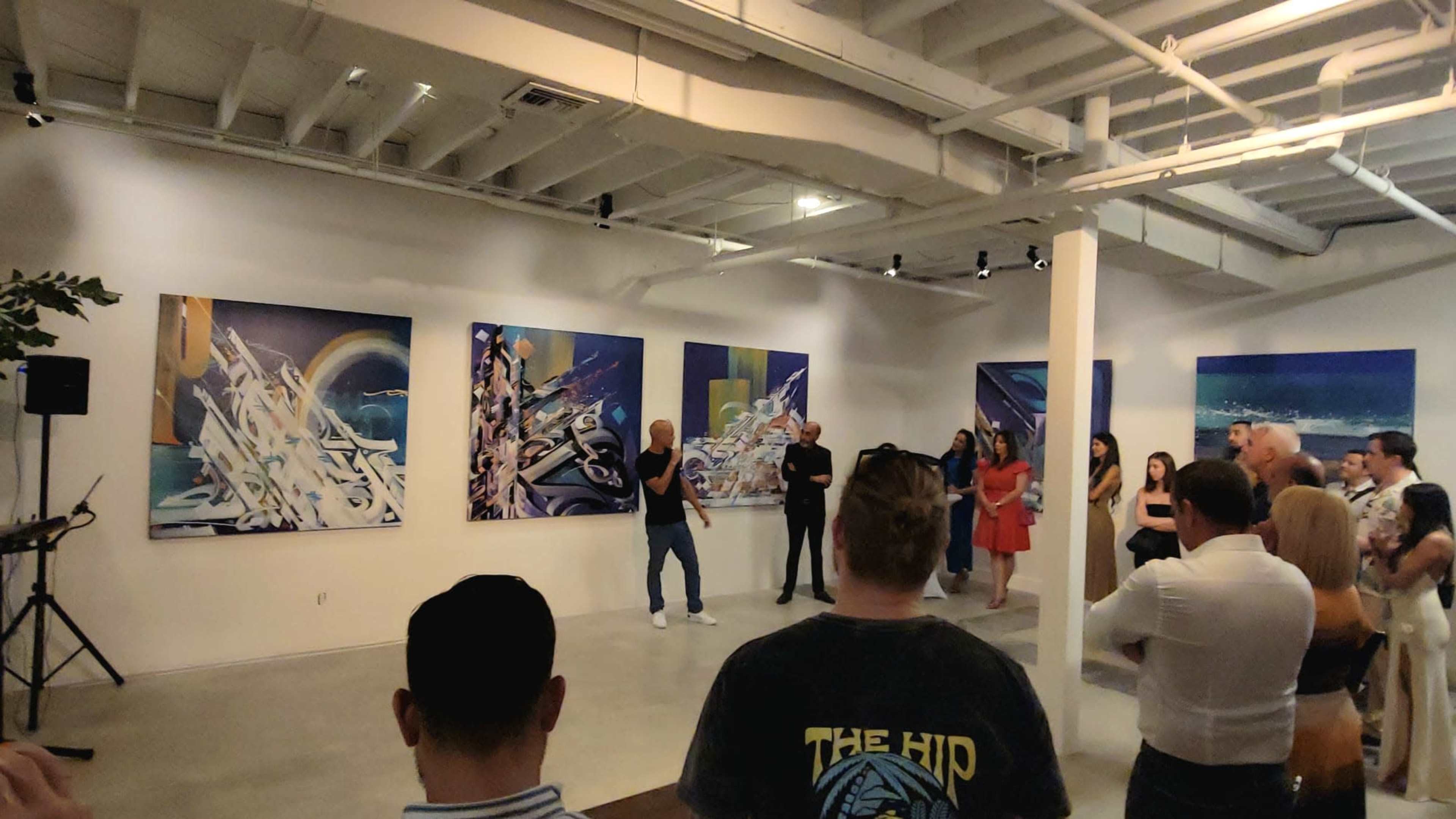 A group of people stands in an art gallery, attentively watching a speaker in front of large contemporary paintings on the walls.