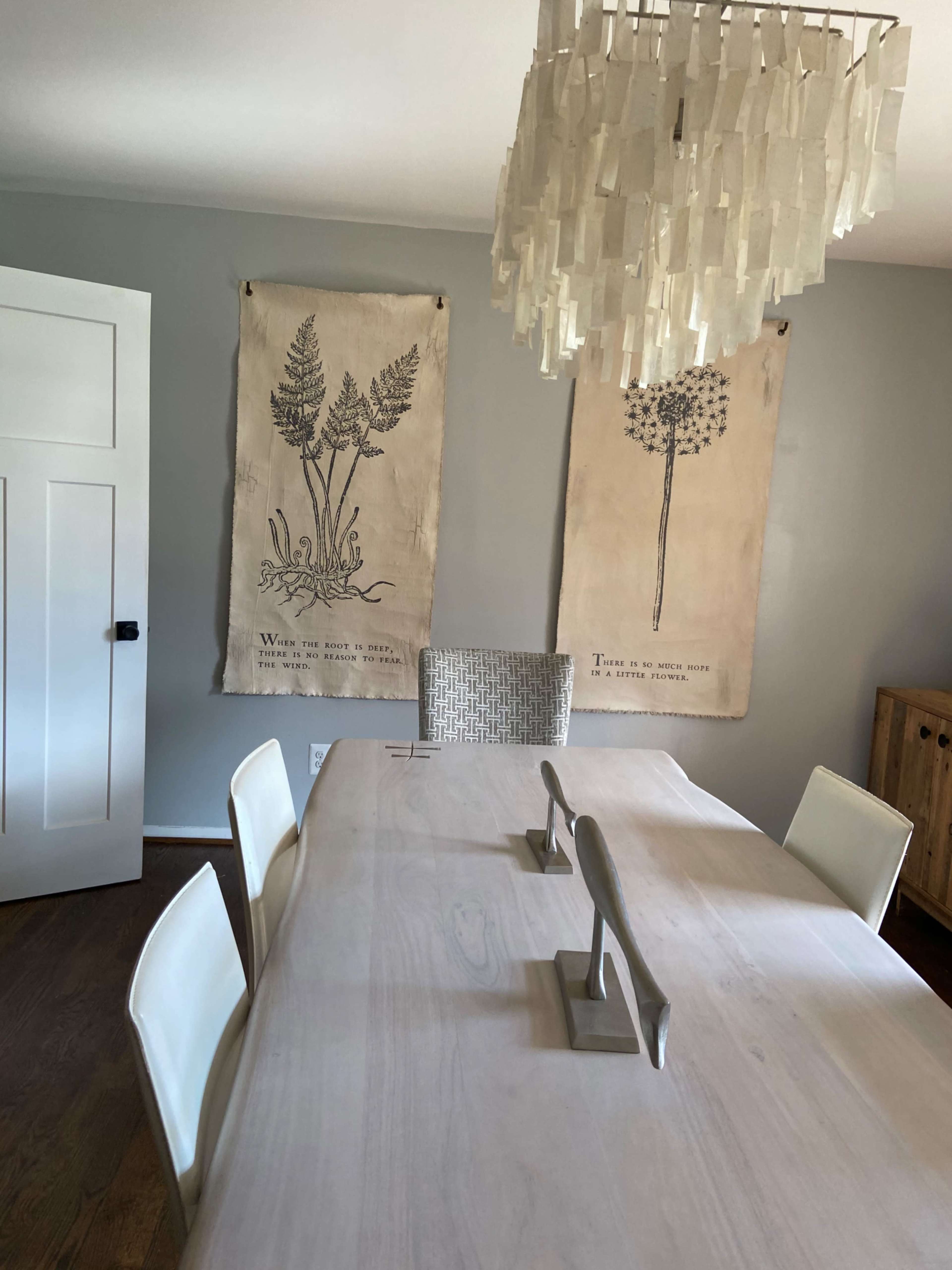 A dining room features a light-colored table surrounded by white chairs, with botanical wall art and a modern chandelier above.