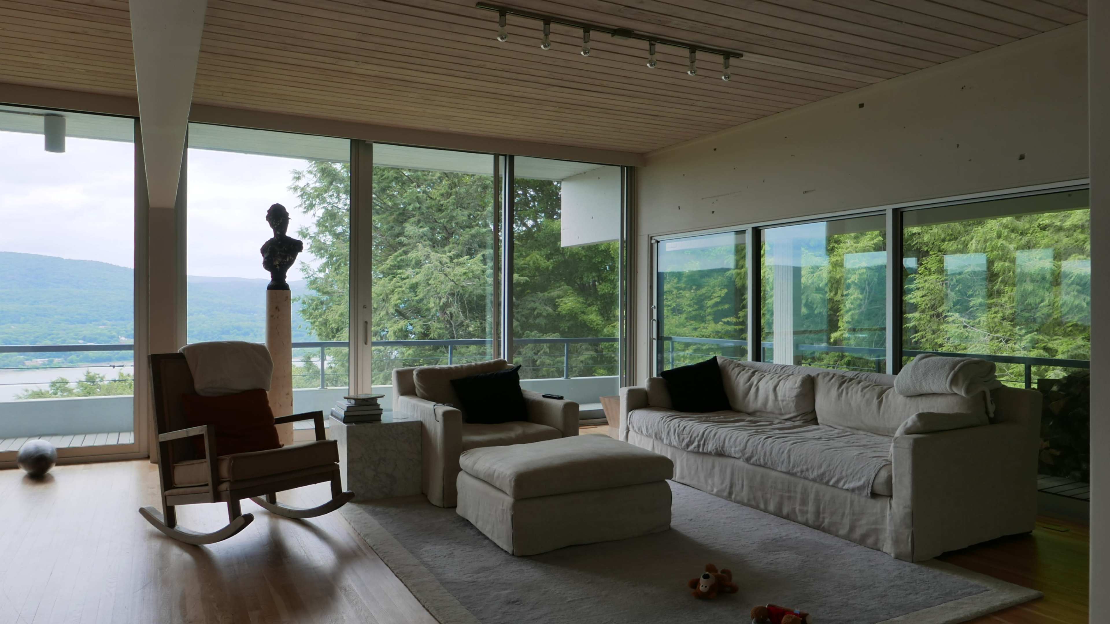 A modern living room with large windows displaying a view of greenery and water, featuring a comfortable seating area, a rocking chair, and a sculpture.