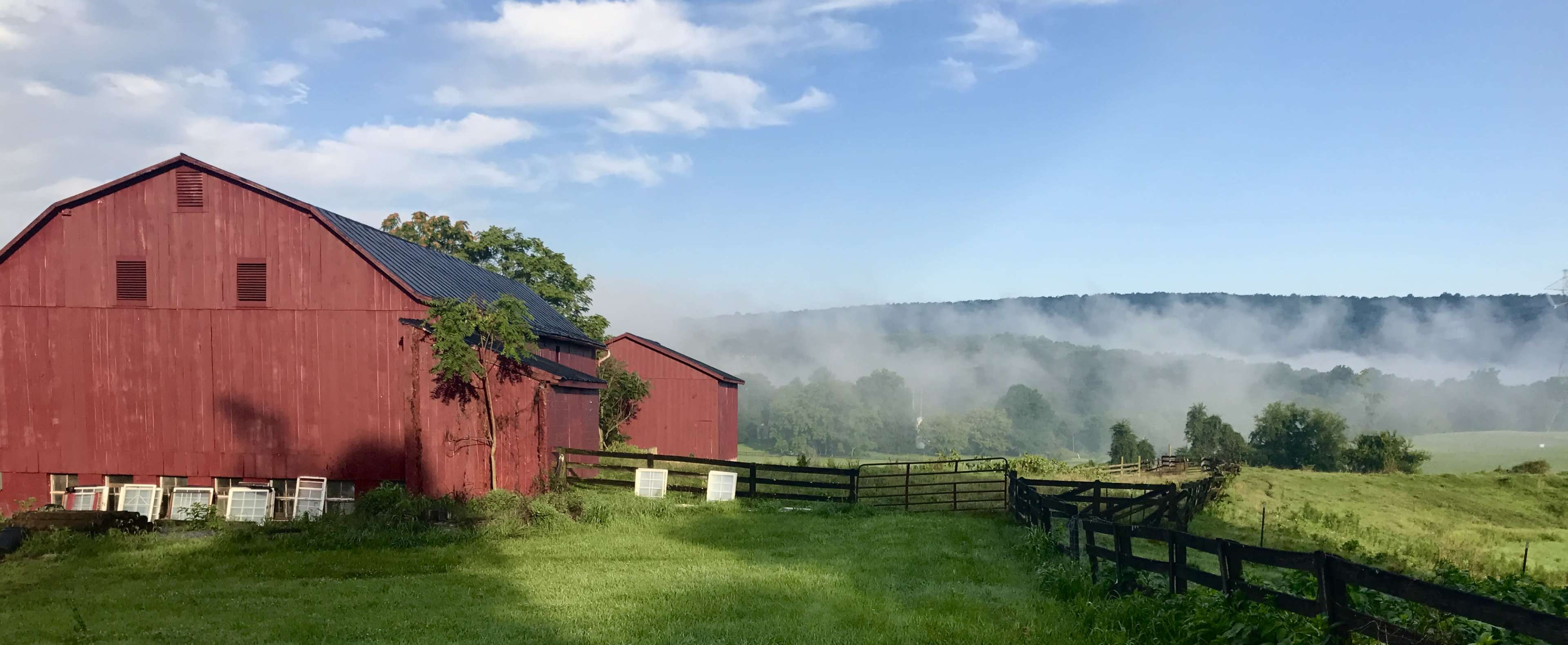 A red barn stands beside a green field, with mist rising from the hills in the background under a clear blue sky.