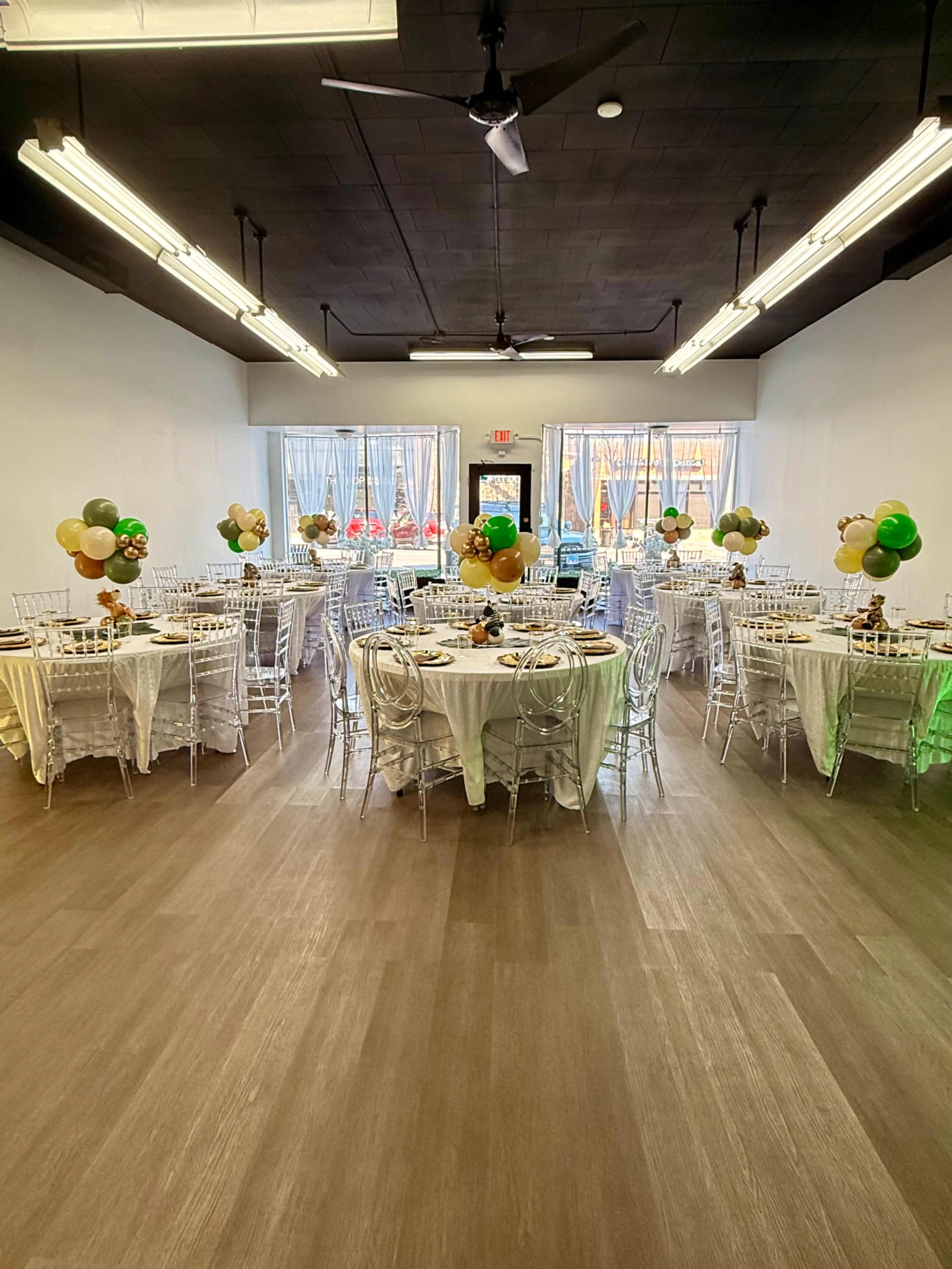 The image shows a well-decorated event space with several round tables set for a gathering, featuring centerpieces of balloons and white tablecloths.