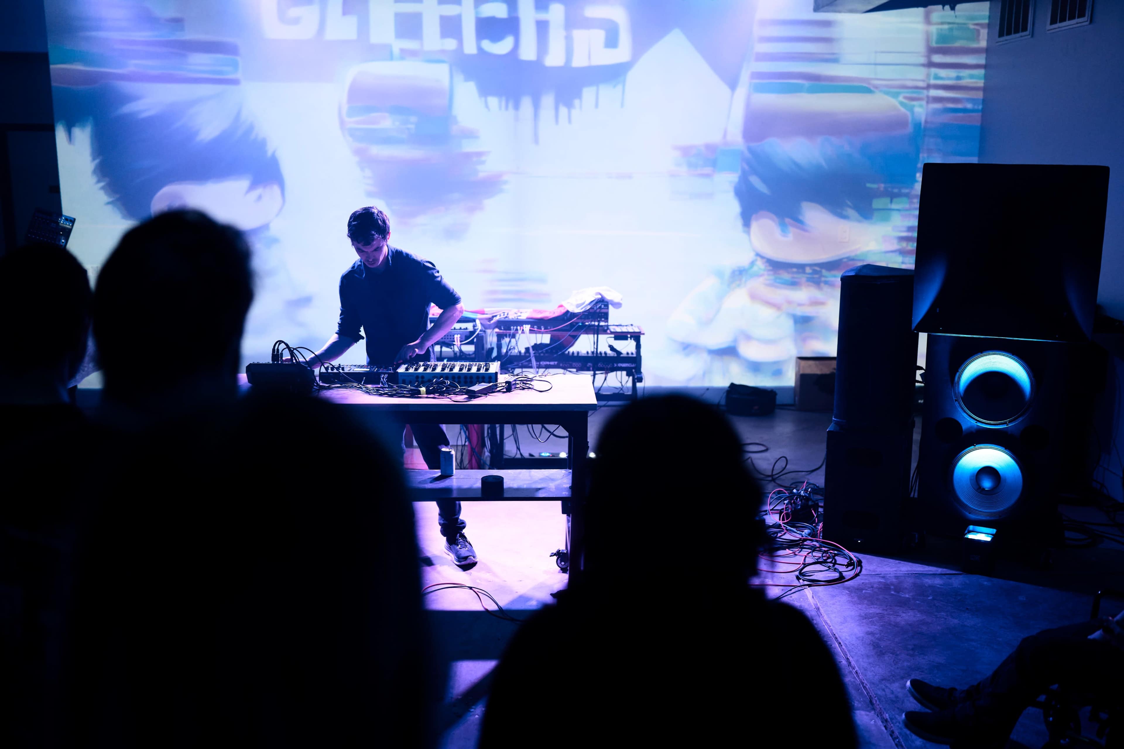 A musician performs on stage in front of an audience while colorful visual projections illuminate the backdrop.