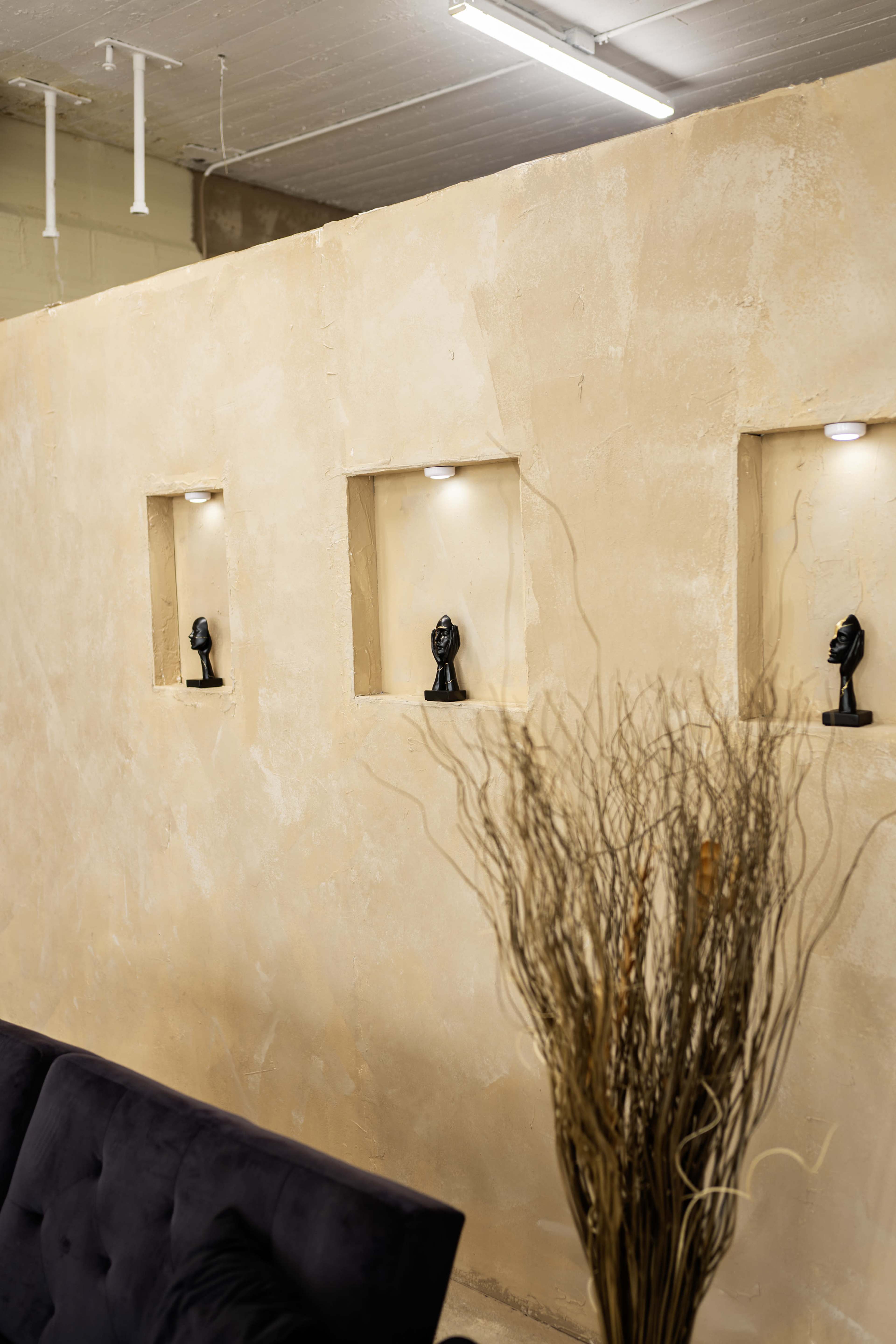 The image shows a textured beige wall with three recessed niches, each containing a small black bust, alongside a dark blue sofa and a tall, dry decorative plant.