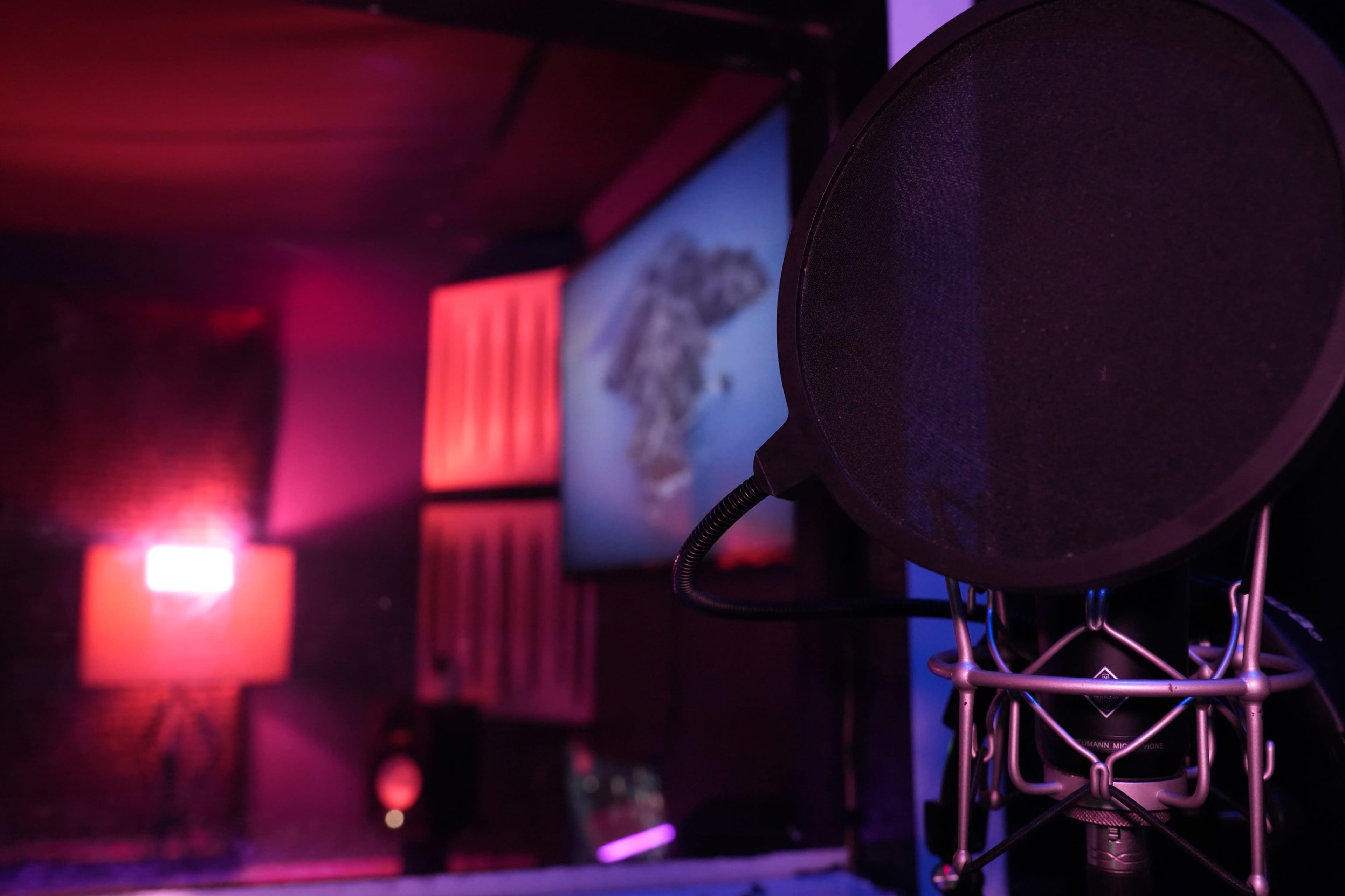 A microphone with a pop filter is positioned in a recording studio with colorful lighting and a screen displaying an image in the background.