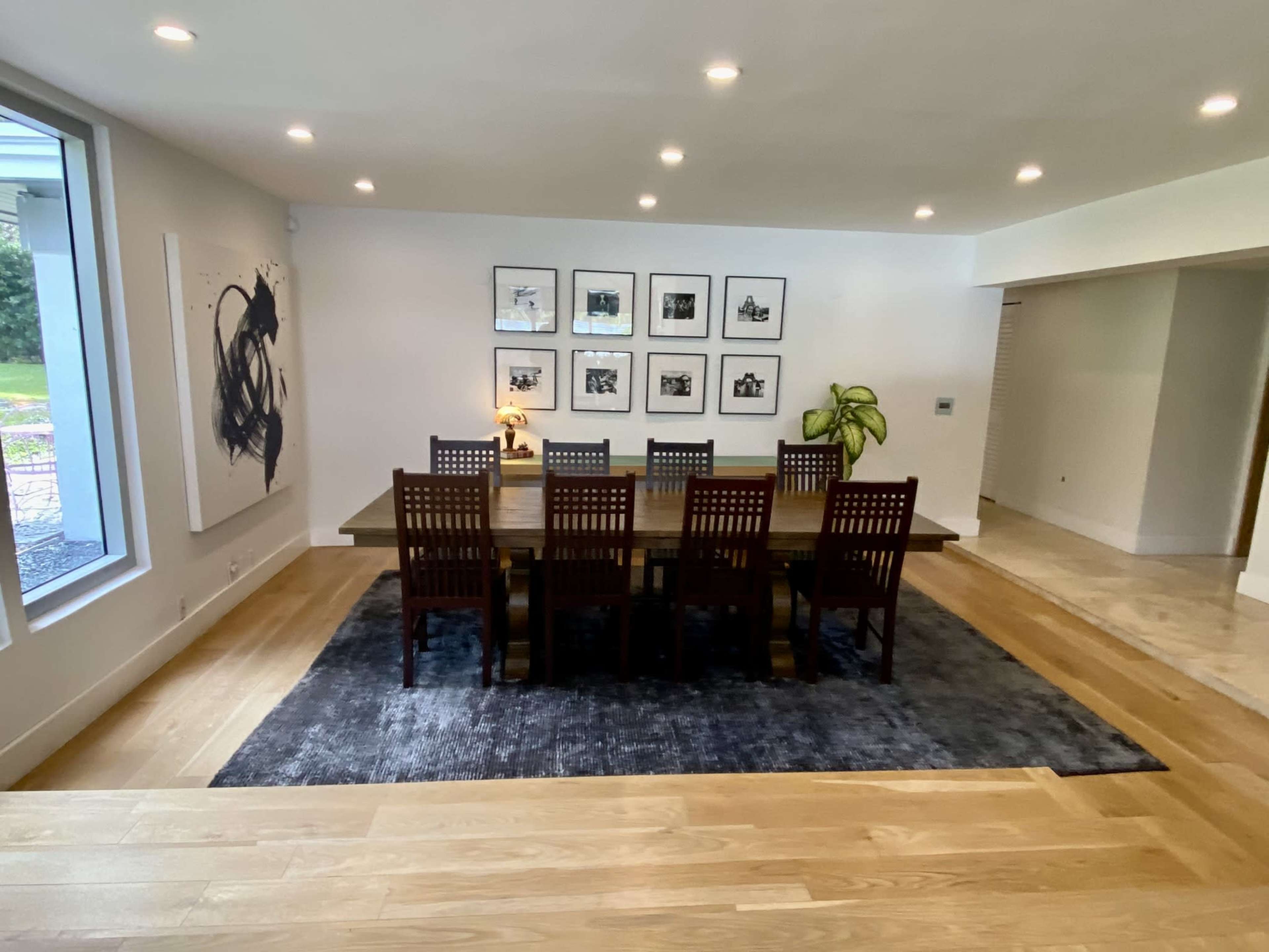 A large dining table with ten wooden chairs is set in a modern dining room featuring framed artwork on the walls and a potted plant nearby.
