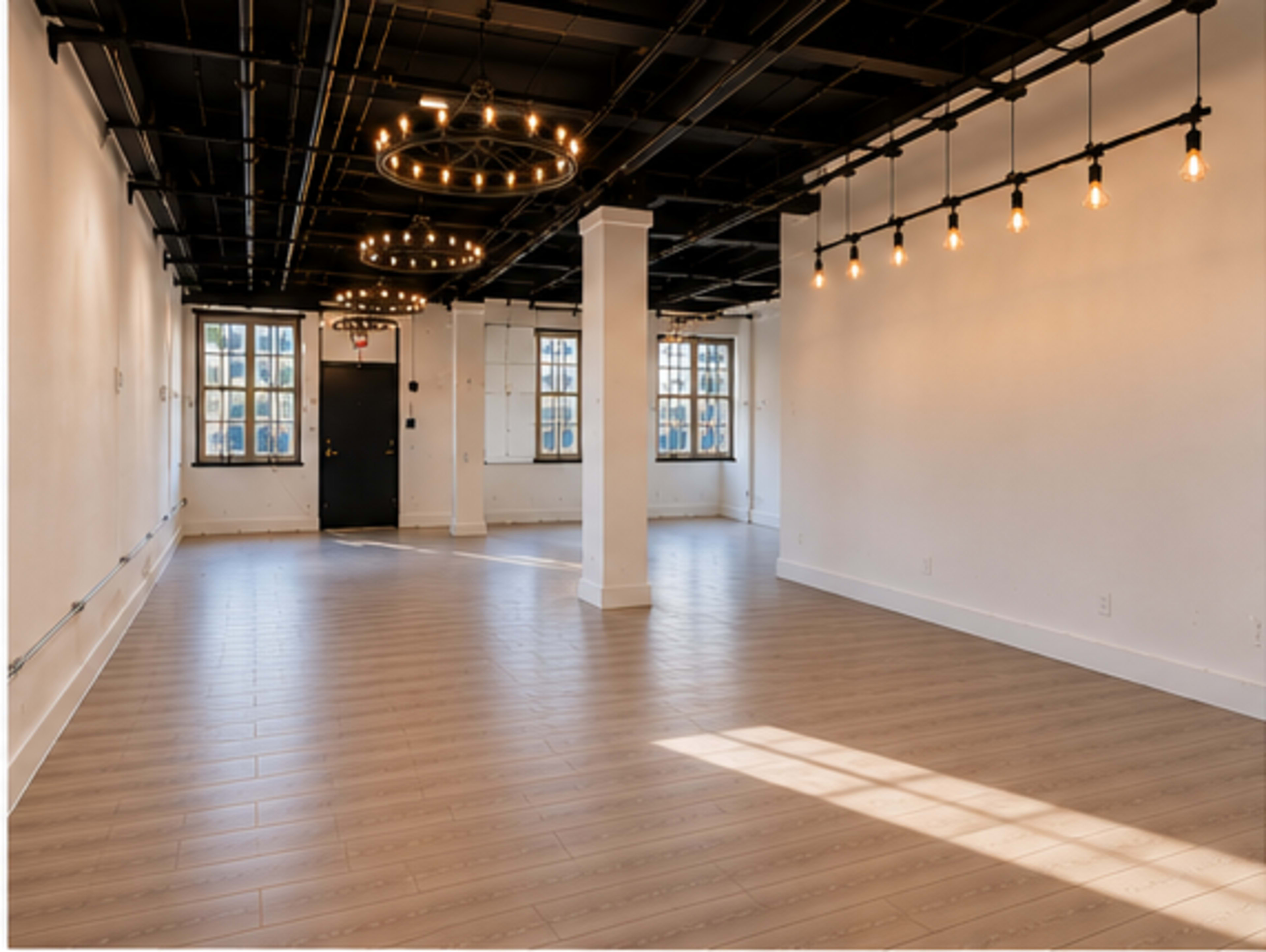 The image shows a spacious, empty room with wooden flooring, large windows, and industrial-style lighting fixtures above.