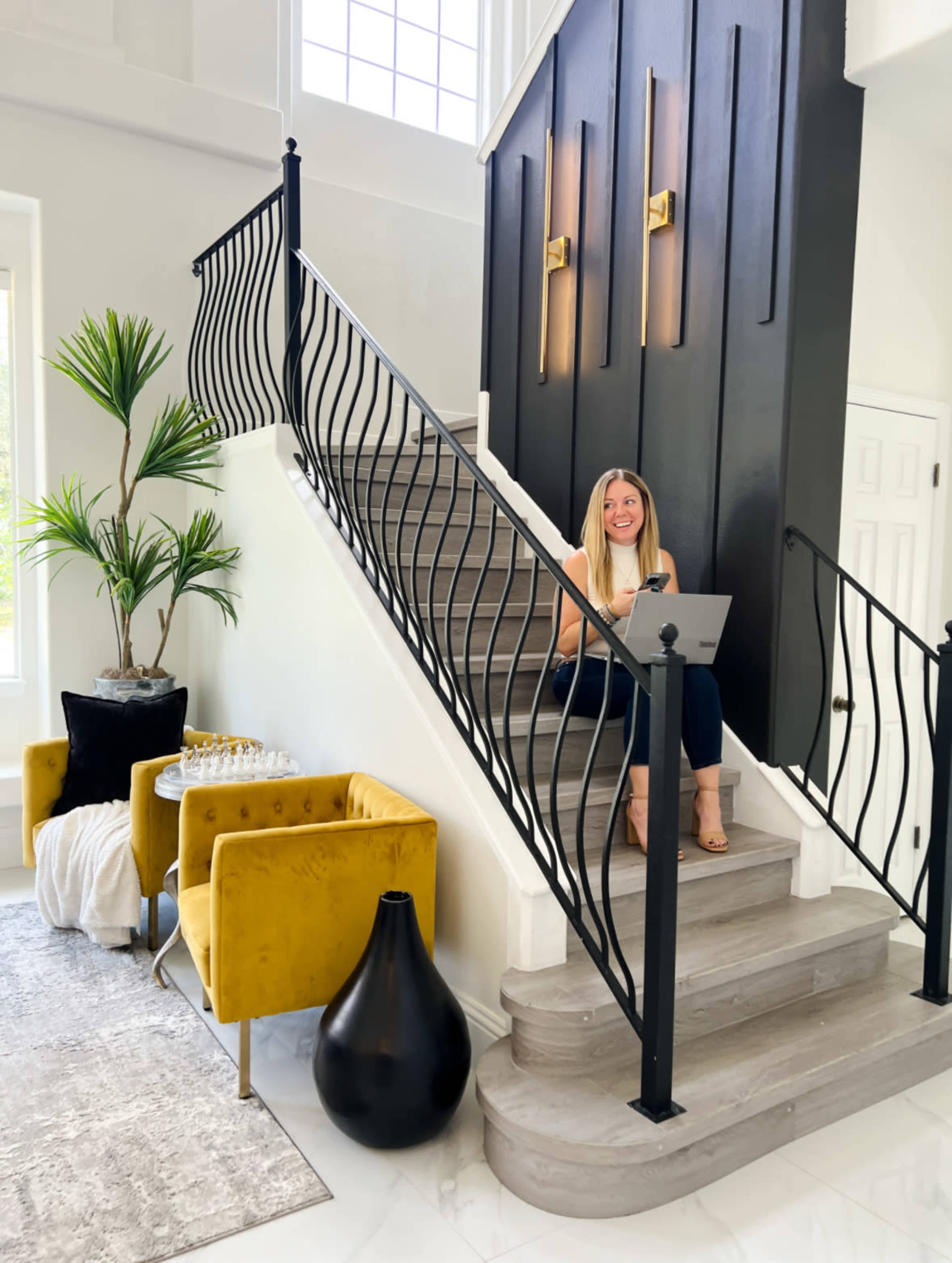 A woman sits on a stairway while working on a laptop, surrounded by decorative plants and modern furniture.